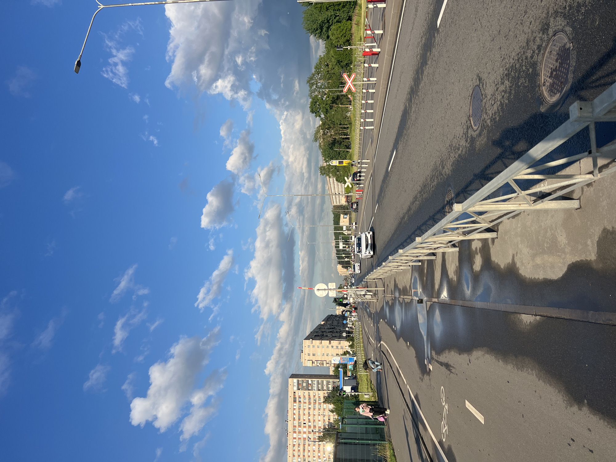 The image shows a wide paved road in an urban environment. On the left side of the road, there is a separate lane marked for bicycles, with a white bicycle symbol painted on it. This lane appears to be wet in patches, with reflections of the sky and clouds visible in the puddles. A person in a pink jacket and dark pants is walking on the sidewalk next to the bike lane, pulling a suitcase. Another person is kneeling on the bike lane, possibly tying a shoelace. In the background on the left, there are several multi-story apartment buildings. The main road has multiple lanes for vehicles, with cars driving in the distance. A white metal fence runs along the right side of the road, separating it from a grassy area and trees. Several traffic lights and road signs are visible. In the far distance, there are more buildings and trees under a bright blue sky with scattered white clouds. The overall lighting suggests it is late afternoon or early evening.