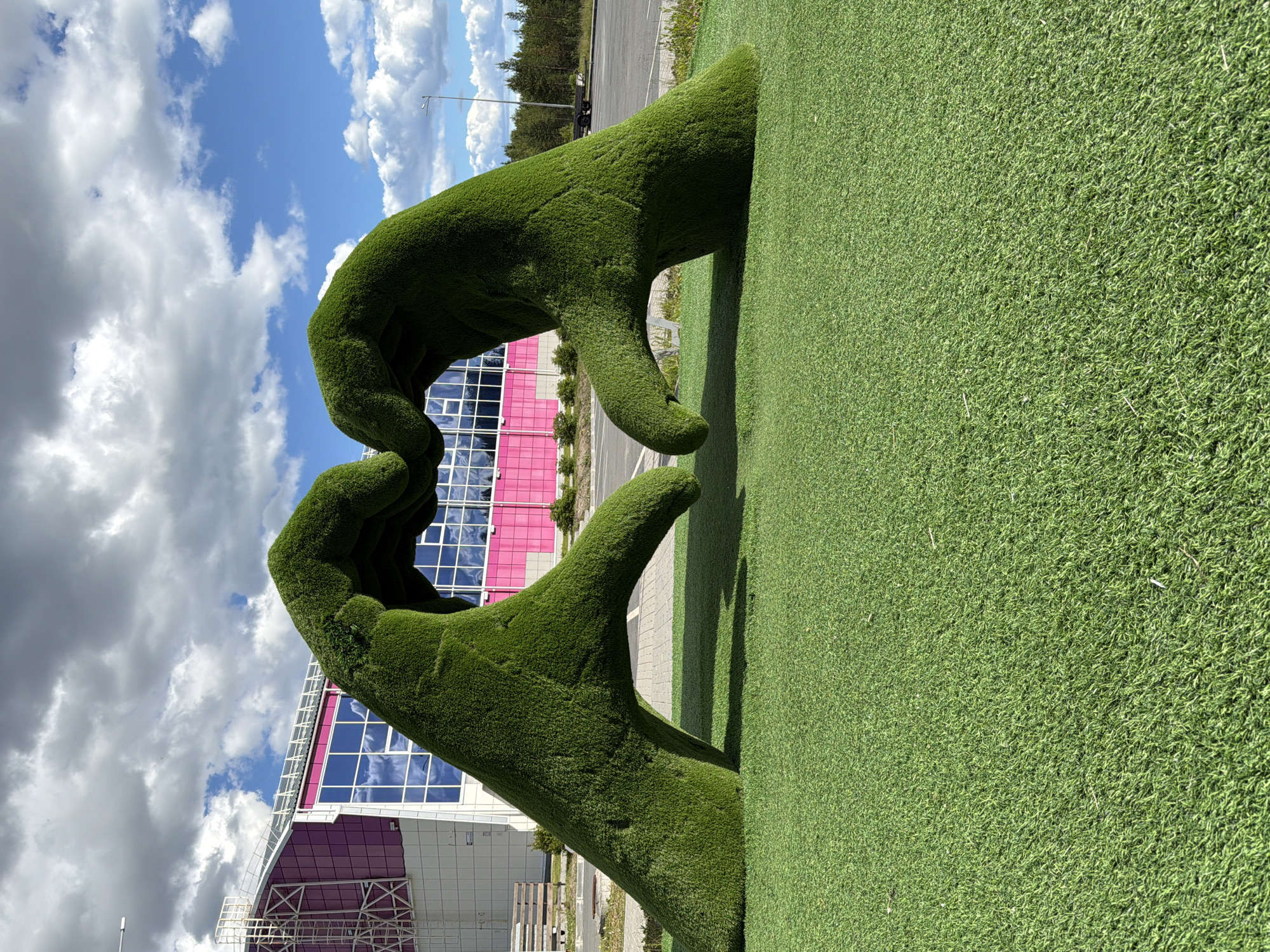 Two large sculptures of hands made of green artificial grass are positioned on a grassy field, forming a heart shape. The hands are angled inwards, with the fingertips touching to create the top of the heart. Behind the sculptures, a building with a pink and white facade and blue windows is visible. The sky is partly cloudy with blue patches. The location is the city of Polarnie Zori, Russia, as indicated by the metadata. The scene is outdoors, with the sculptures and building set against a natural backdrop of trees in the distance and a cloudy sky.