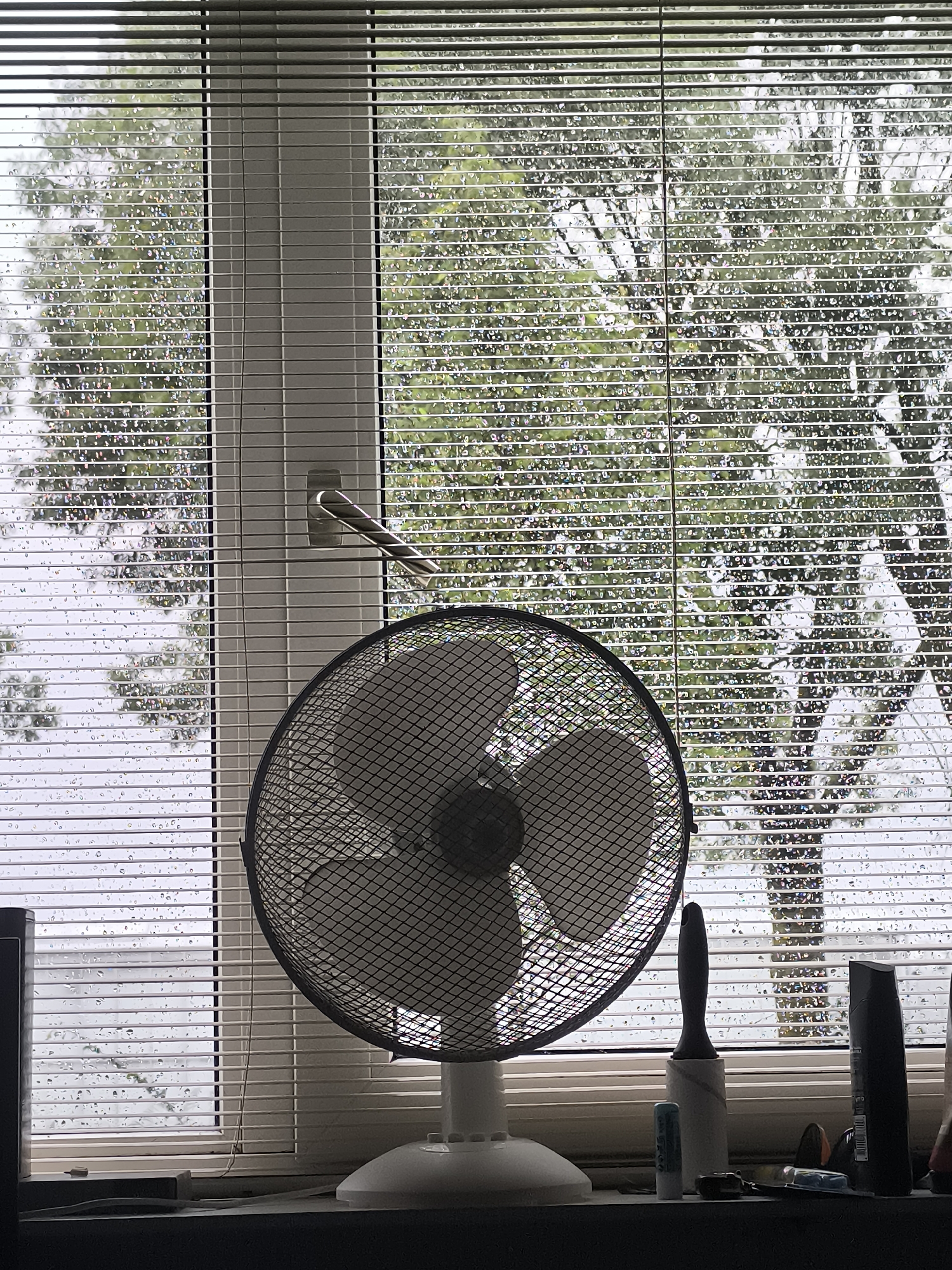 The image captures a close-up view of a window with blinds and raindrops, likely taken from an interior. A desk fan, white with a black grille, is prominently positioned in the foreground, partially obscuring the view outside. The window appears to be a standard residential or office window. The blinds are horizontal and adjustable, allowing some light to filter through. The exterior scene visible through the blinds shows trees with green foliage, suggesting a temperate climate. The presence of raindrops on the windowpane indicates recent or ongoing precipitation. A silver-colored window handle is visible on the right side of the window frame, positioned between the blinds. On the windowsill below the fan, several items are scattered, including a lint roller, a small blue and white object, and a dark-colored bottle, possibly a toiletry item. The overall impression is one of a slightly overcast and damp day, viewed from indoors. The location is specified as Almere, Netherlands.