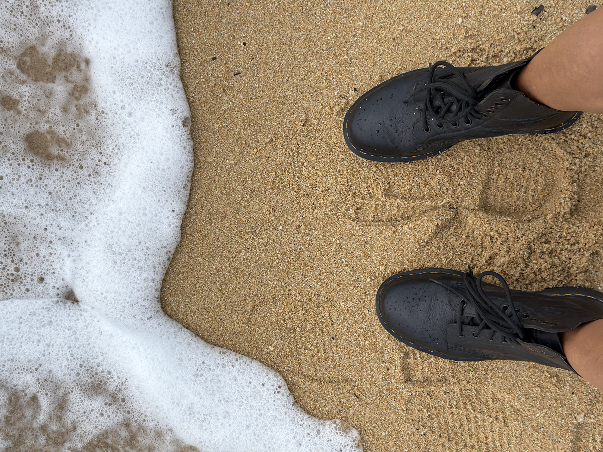 A pair of black leather boots stand on a sandy beach, with foamy white water lapping at the toes of the boots. The sand is a light brown color and appears damp. The boots are laced up and have yellow stitching. The scene is photographed from a high angle, looking down at the boots and the beach. The location is the beach in Ko Samui District, Thailand.