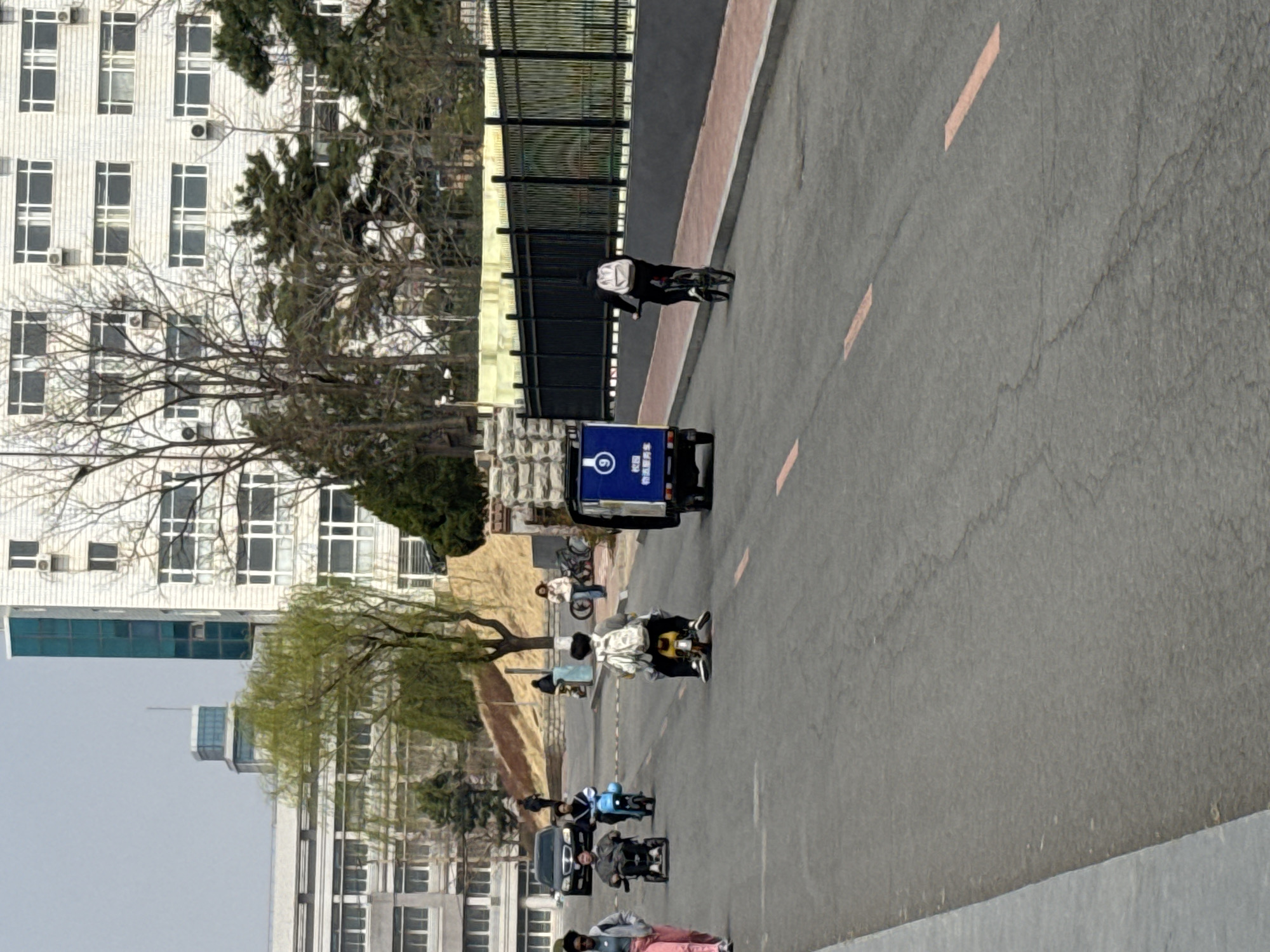 The image depicts a daytime scene on a paved road within what appears to be an urban or university campus environment in Zhongtun, China. The road features light orange dashed lines and a darker sidewalk strip alongside it.
In the middle ground, several individuals are navigating the road, predominantly riding electric scooters or bicycles. One person, dressed in dark clothing with a white backpack, is riding a bicycle near a black metal fence on the right side of the road. Further along the road, a blue three-wheeled electric vehicle, likely a cargo or delivery transport, is prominent. Its side displays a blue circular logo with the number "9" and Chinese text that reads "校园" (campus) and "物资存放" (material/goods storage). Other individuals on electric scooters and bicycles are seen further down the road, and a dark-colored car is partially visible in the foreground.
The background is dominated by several multi-story buildings, mostly white or light-colored, featuring numerous windows and visible air conditioning units, characteristic of residential or institutional architecture. Trees are scattered across the scene; some are bare, suggesting an early spring or late winter, while others exhibit fresh green foliage. The sky is clear and bright, indicating fair weather. The scene conveys a sense of routine daily activity and movement within a local setting.