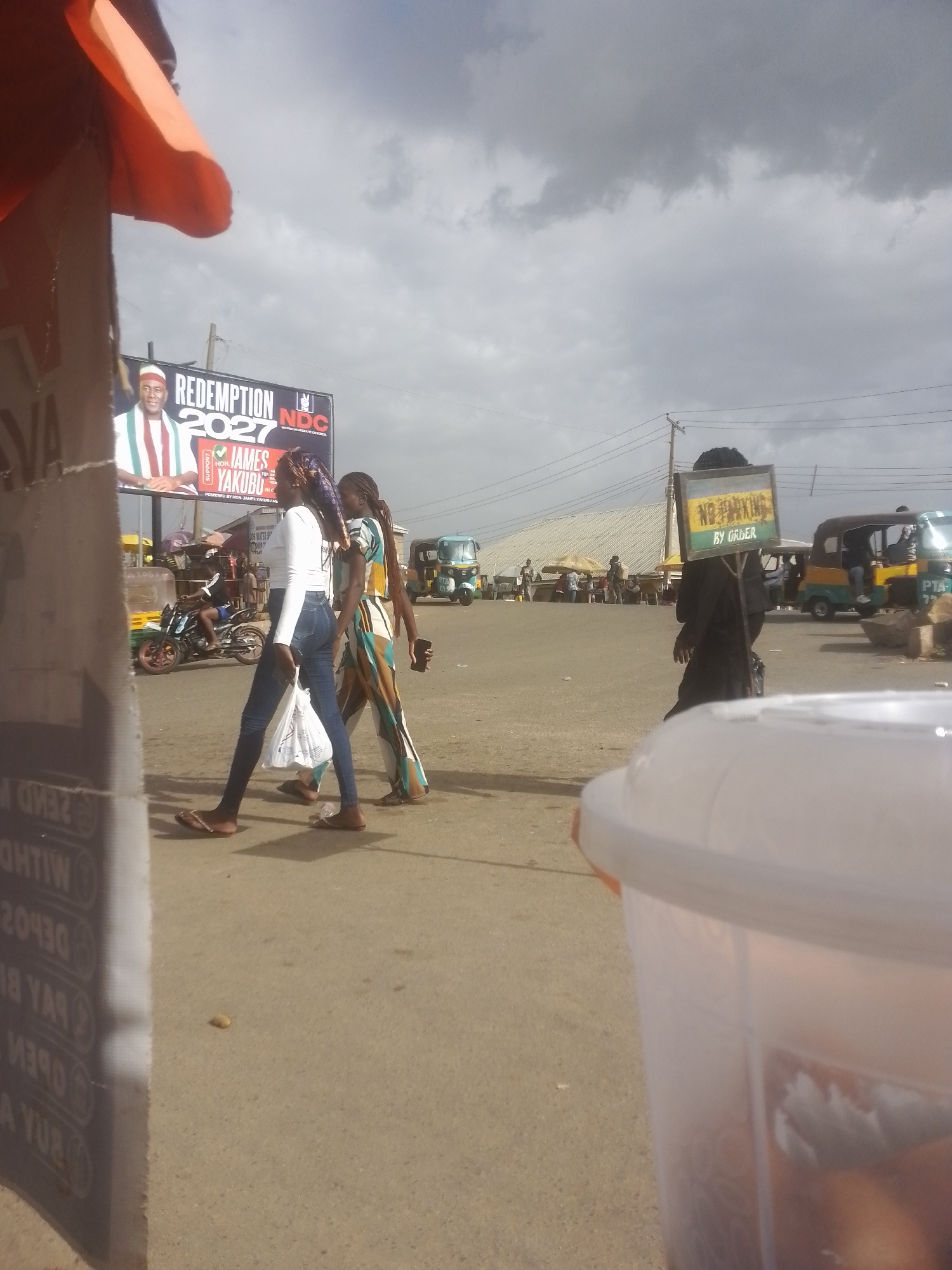 A man in a red and white striped hat and a green, white, and red striped sash is pictured on a billboard in the background. Text on the billboard reads "REDEMPTION 2027" and "HON. JAMES YAKUBU". Two women walk past the billboard. The woman on the left wears a white long-sleeved shirt and blue jeans, while the woman on the right wears a multi-colored striped dress. In the background, a man on a motorcycle drives past, and several tuk-tuks are parked. A person carries a sign that reads "NO PARKING BY ORDER". The scene appears to be a street or market area with a cloudy sky.
