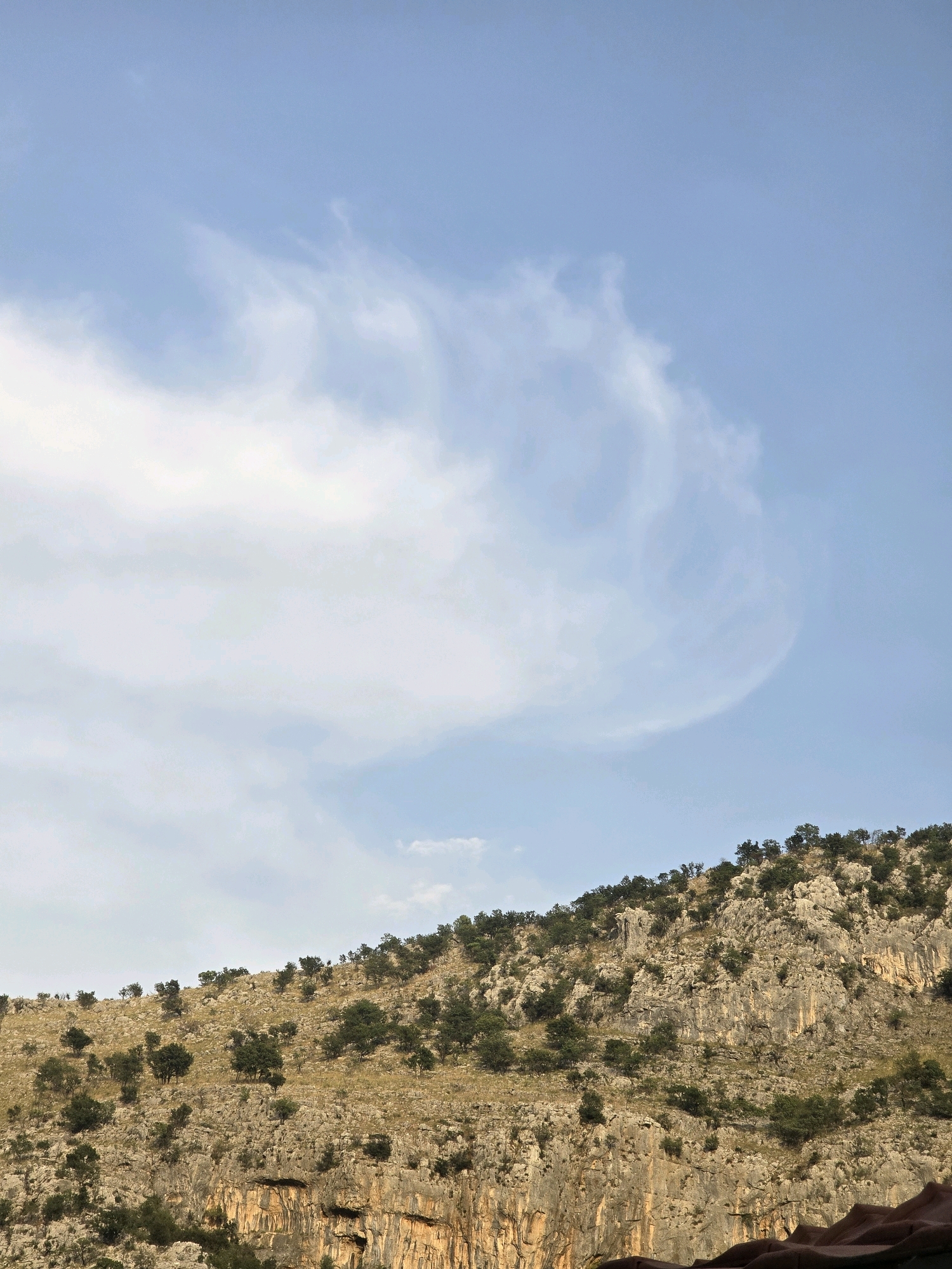 The image depicts a rocky, tree-covered mountainside under a bright blue sky with wispy clouds. The mountain is steep and appears to be part of a rugged landscape. The trees are scattered across the slopes, with some appearing denser in certain areas. The terrain is characterized by rocky outcrops and crevices. The lighting suggests it is daytime, likely with sunlight illuminating the scene. The photograph's composition focuses on the natural environment of the mountain. There are no people or man-made structures clearly visible in the image. While the description focuses on the visual elements, it does not specify a location within the city of Podgorica, Montenegro.