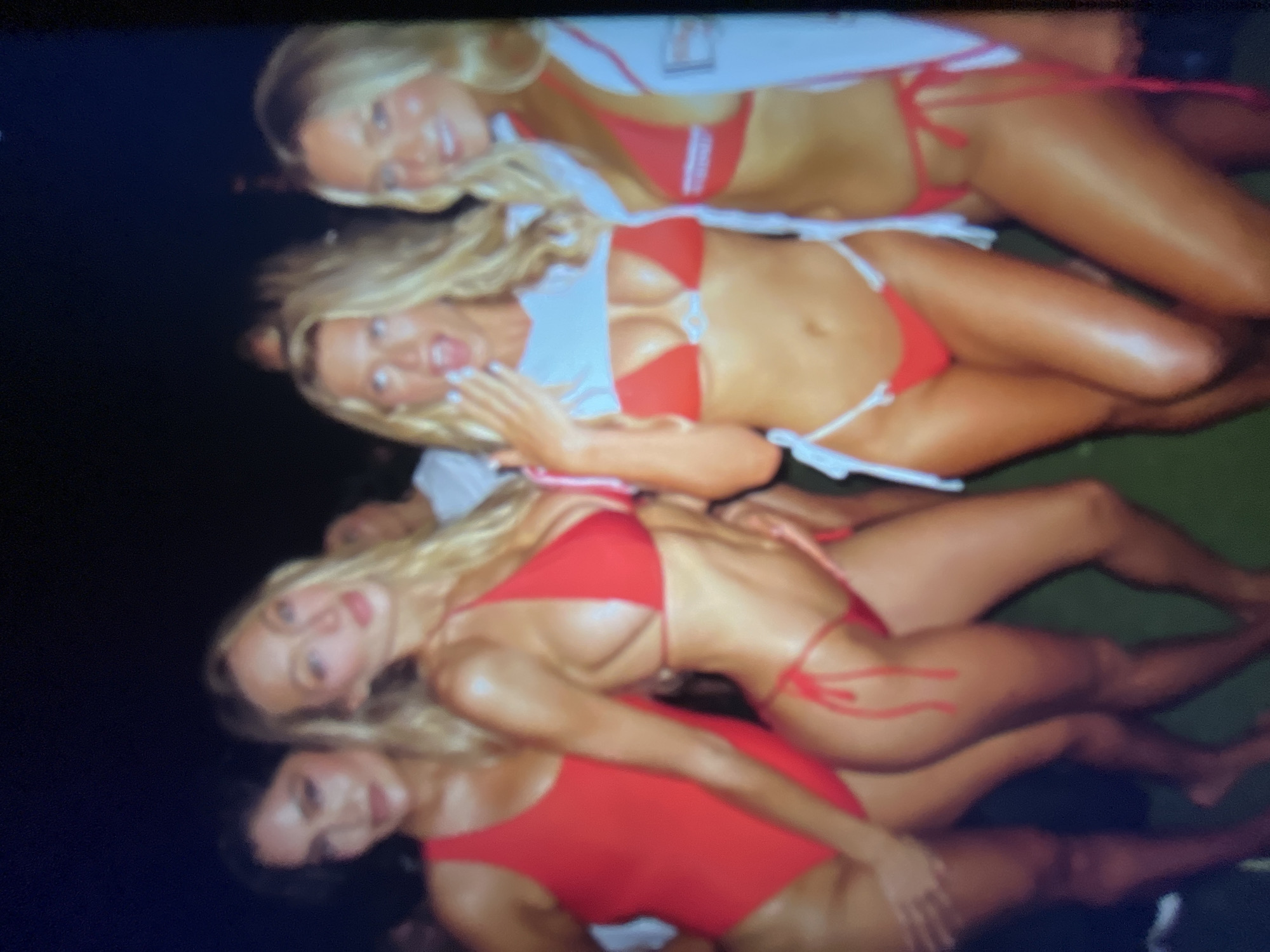 A group of four young women, all wearing red swimwear, are posing for a photograph at night. The woman on the far left is wearing a red one-piece swimsuit, while the other three are in bikinis. The woman second from the left is wearing a red bikini with a white trim. The woman in the center is wearing a red bikini with a white, cropped top over it, and her mouth is open. The woman on the far right is wearing a red bikini with a white trim and a white jacket with red accents. They are standing on a green surface, possibly grass or Astroturf. The background is dark, suggesting it is nighttime. No specific location in the city of Sofia, Bulgaria, can be identified from the image.