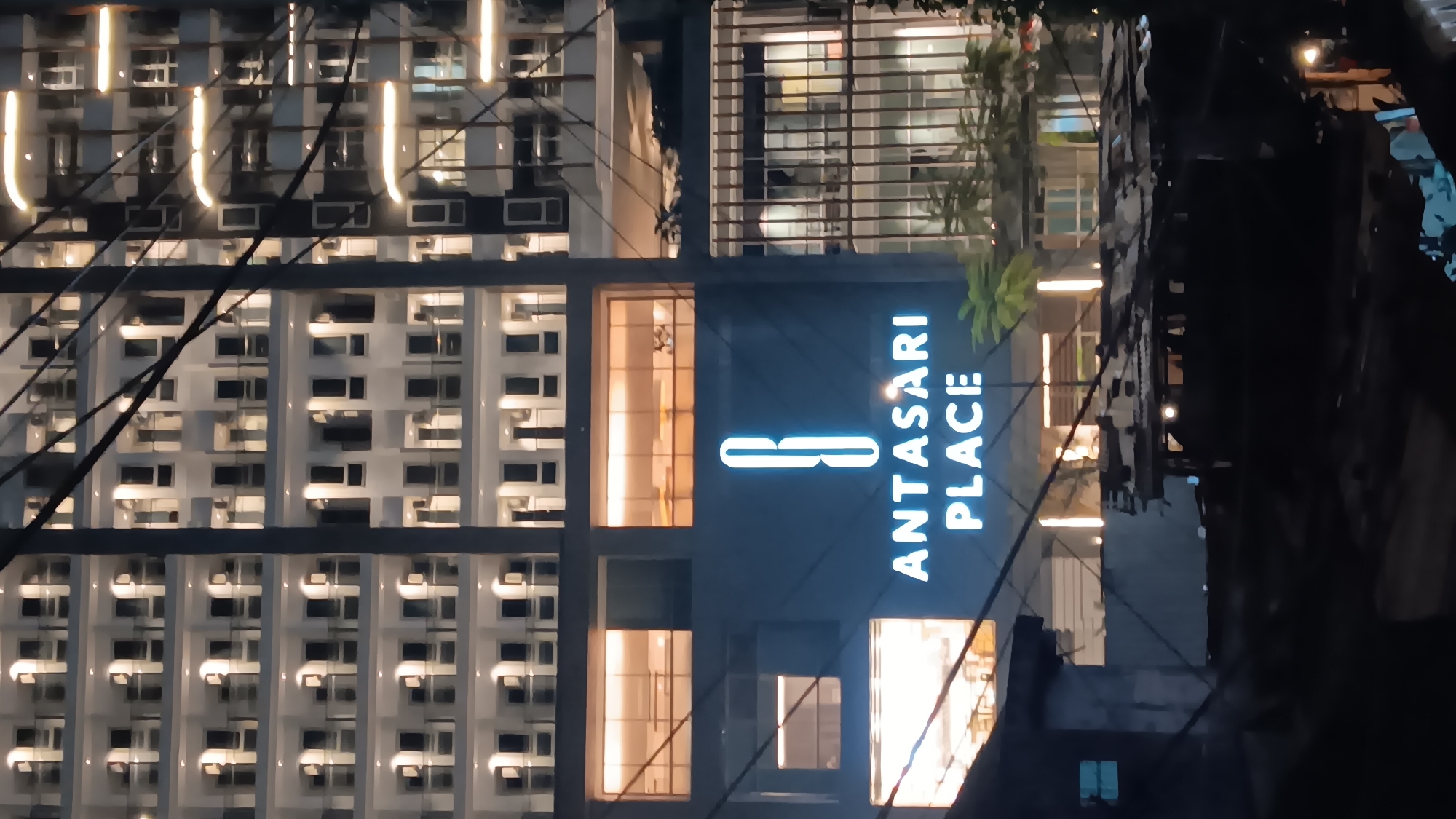 The image shows a nighttime view of a modern building facade with the signage "ANTASARI PLACE" illuminated in bright white letters. The building is multi-story, with numerous windows and balconies visible, many of which are lit from within. The facade appears to be a dark color, possibly blue or grey, contrasting with the bright signage. The building is located in the Kebayoran Baru district of South Jakarta, Indonesia. In the foreground, there are some darker, more rudimentary structures with corrugated metal roofs, suggesting a less developed area juxtaposed with the modern building. Several dark wires or cables are strung across the upper portion of the image, obscuring parts of the building. The lighting is primarily artificial, creating strong contrasts between illuminated areas and shadows. There are no people visible in the image.