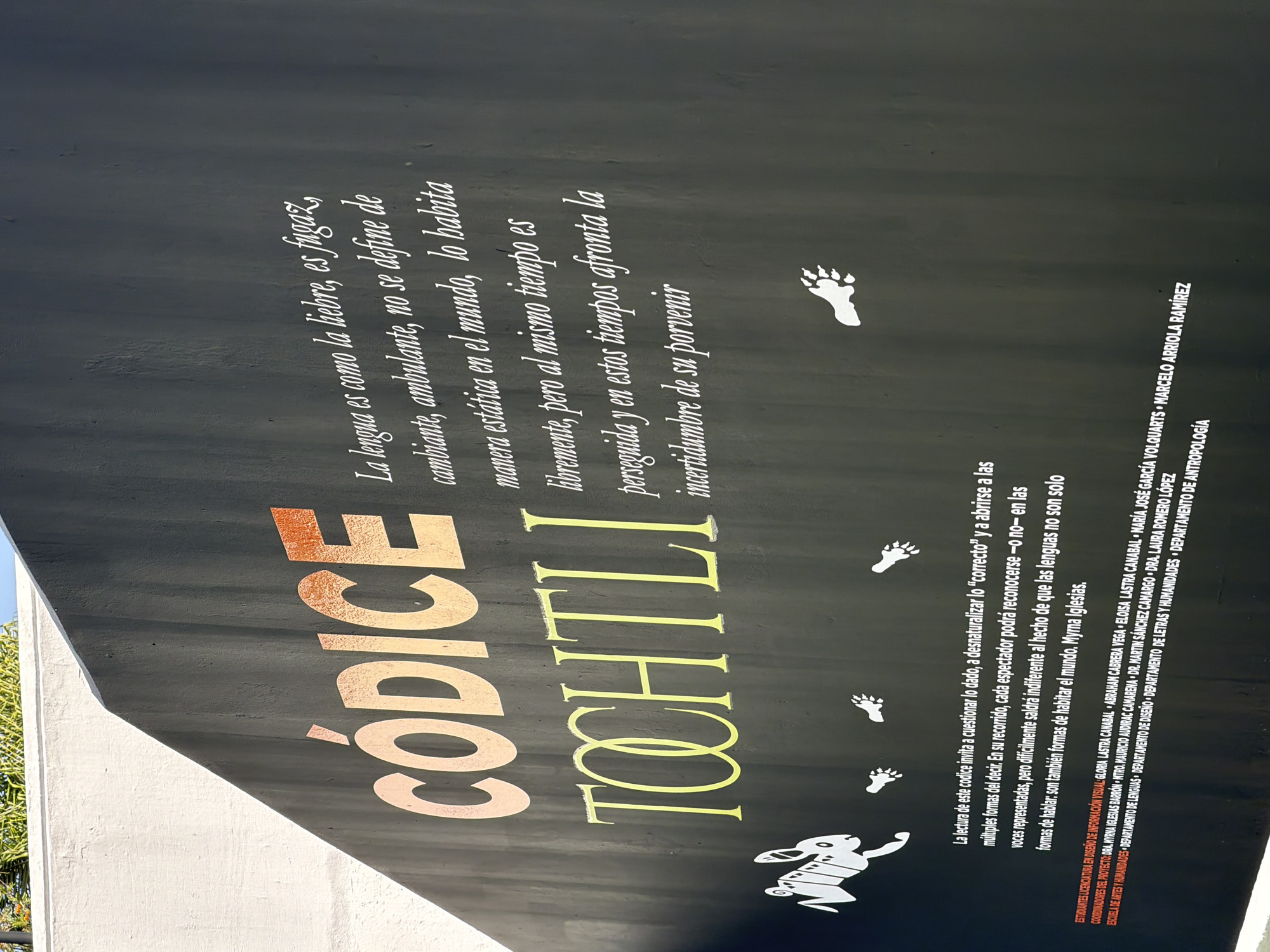 Here is a description of the image:
The image shows a close-up of a black informational sign or display. The main text on the sign reads "CÓDICE TOCHTLI," with "CÓDICE" in a copper-colored, large font and "TOCHTLI" in a yellowish font. To the right of the text, there is a longer paragraph describing the nature of language, comparing it to a hare and stating that it is not static but lives freely.
Below this, there's a white illustration of a hare standing upright, with several white paw prints scattered upwards. Underneath these illustrations and texts, another paragraph is present, discussing the interpretation of the codex. It mentions that the reader is invited to question what is given and to open up to the multiple forms of saying, and it concludes by noting that languages are not just ways of speaking but also ways of inhabiting the world.
At the bottom of the sign, there is smaller text listing names and departments involved in the project, including the coordinators, students in design of information visualization, and the departments of languages, design, humanities, and anthropology.
The setting is likely outdoors, given the natural lighting and possible vegetation visible in the background. The sign is mounted on a structure, and the overall atmosphere suggests an educational or cultural exhibit, possibly in a public space in Unknown, Unknown.