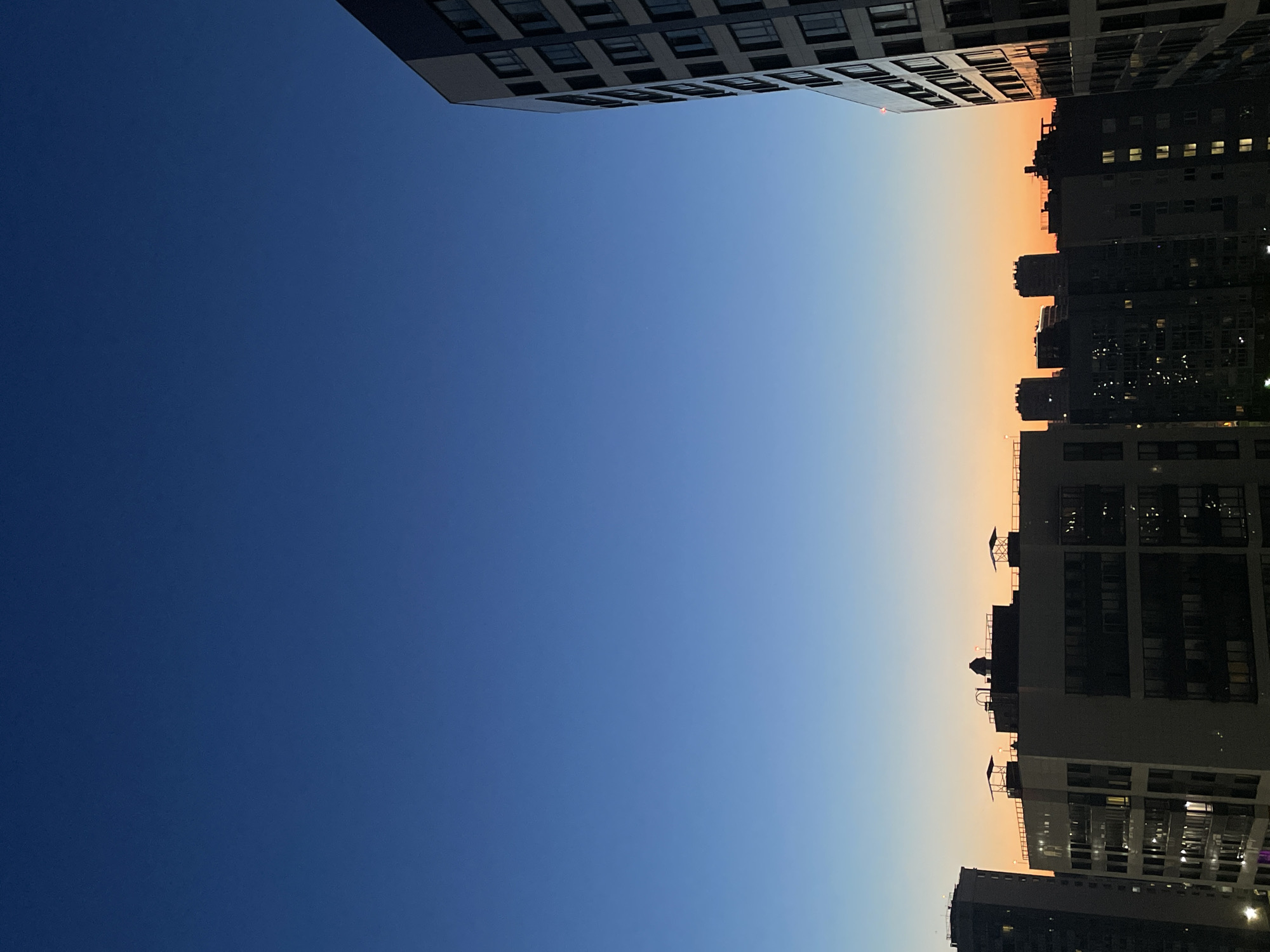 The image captures a twilight view of Sinyavino, Russia, showcasing a cityscape against a gradient sky transitioning from deep blue overhead to a warm orange hue near the horizon. Silhouetted apartment buildings dominate the lower portion of the frame, their windows reflecting the fading light and the glow of interior illumination. The architectural style appears modern, with several multi-story structures of varying designs visible. One prominent building on the right side of the image is brightly lit, revealing a grid of windows. The sky is clear, with no visible clouds or celestial bodies, suggesting the very early or late stages of twilight. The overall atmosphere is serene and urban, with the buildings standing as testament to human presence in the landscape. There are no people or specific actions depicted in the scene.