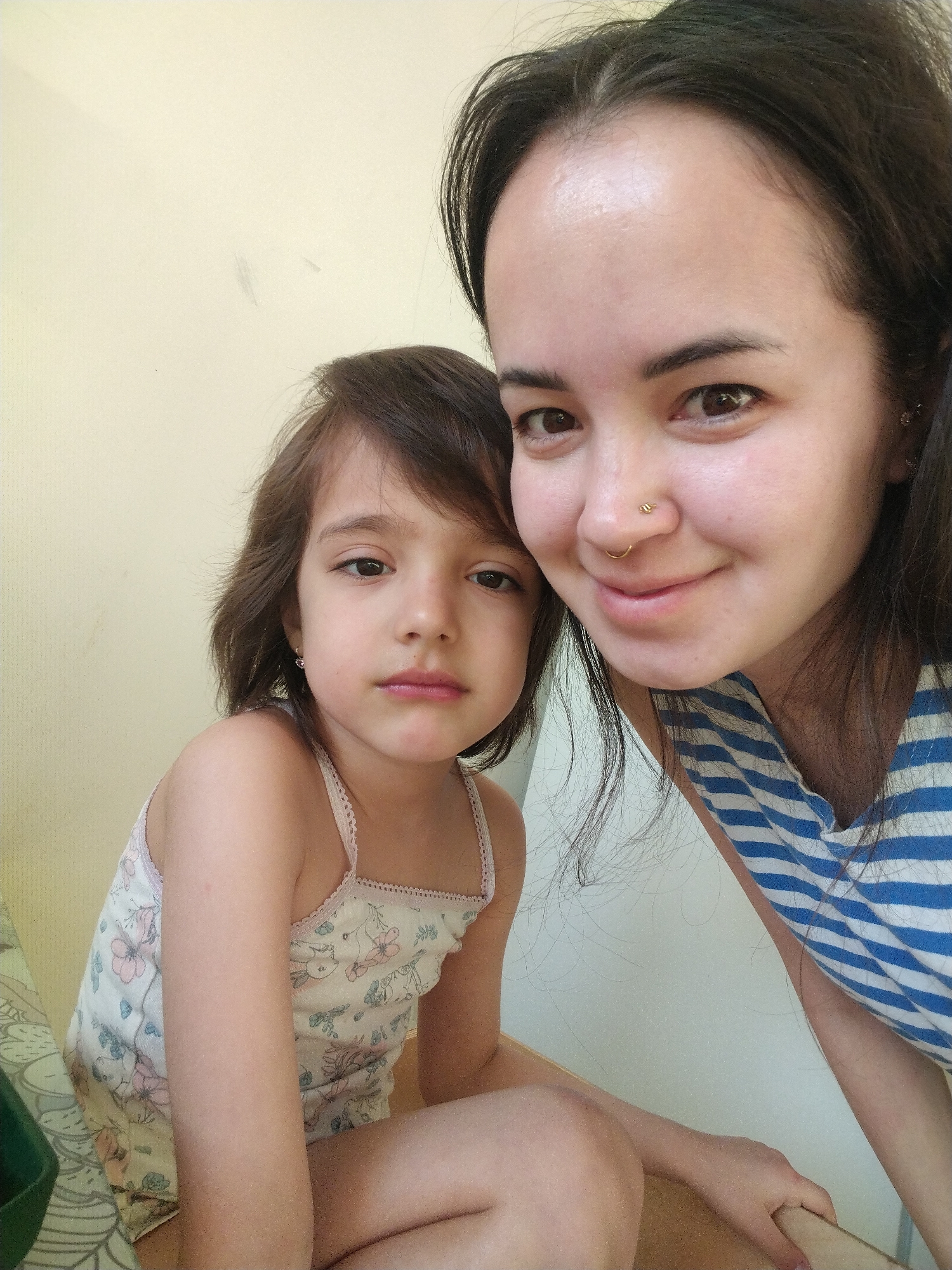 A fair-skinned adult female and a young, fair-skinned child are posing closely for a selfie indoors. The adult female has dark hair, brown eyes, a nose piercing, and is wearing a blue and white striped shirt. The child, who is possibly a young girl, has brown hair, brown eyes, a small earring, and is wearing a light-colored patterned tank top. They are positioned against a light yellow or beige wall. There is no evidence in the image to indicate it was taken in Noviy, Russia.
