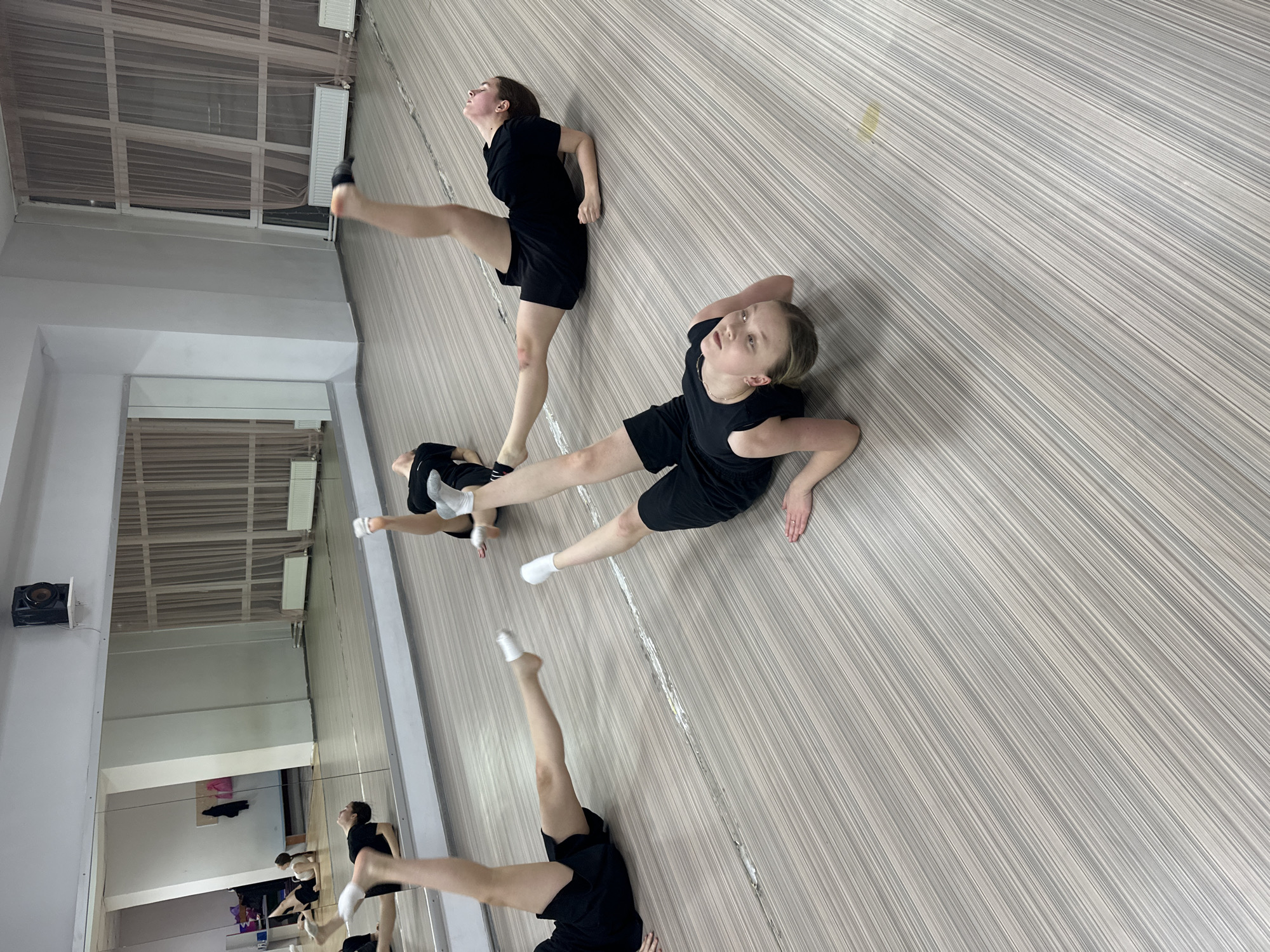 Two young women in black athletic attire are practicing dance or gymnastic poses in a dance studio. One woman is positioned diagonally in the upper right portion of the frame, standing with her left leg extended upwards. The other woman is lying on the floor with her legs spread apart in a straddle position. A large mirror lines the left side of the room, reflecting the dancers and the studio environment. The floor is covered with a striped light and dark colored pattern. The studio appears to be well-lit with white walls, and white ceiling with visible rectangular ceiling lights. The location is Votkinsk, Russia.
