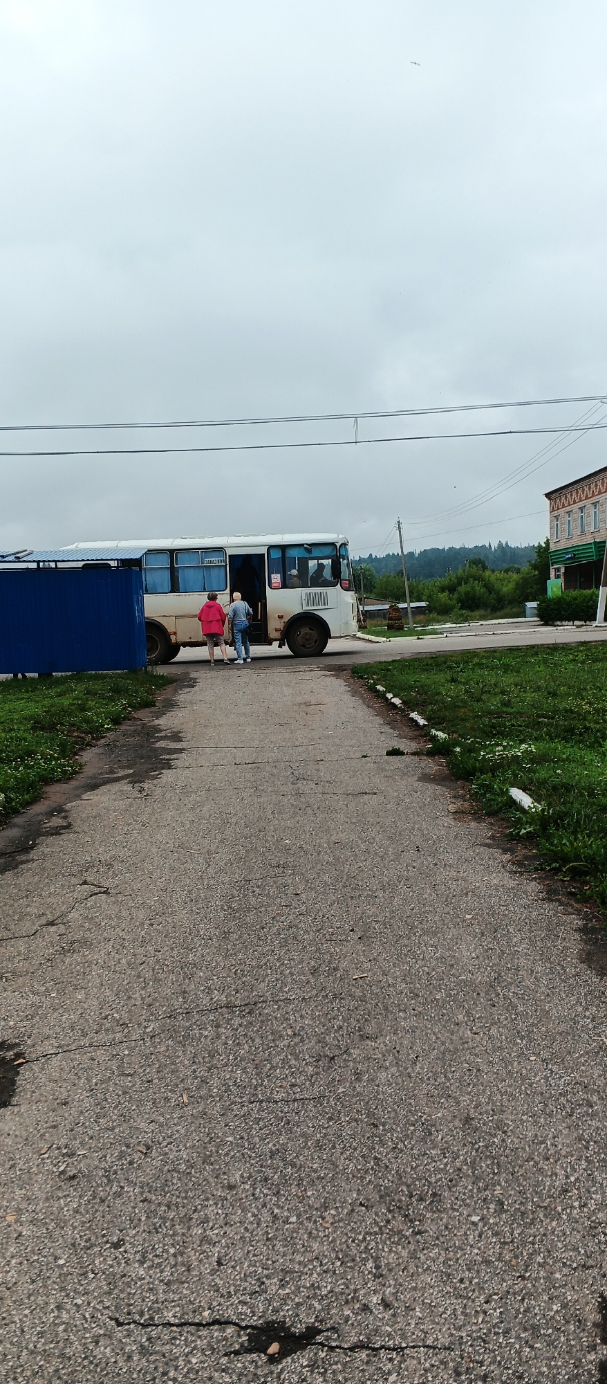 The image depicts a cloudy day scene in the village of Podgornoye, Russia. In the foreground, a cracked asphalt path leads towards a white bus with its door open. Two individuals, one wearing a pink jacket and the other in a blue jacket and jeans, are walking towards the bus. The bus has blue-tinted windows, and text is visible on the side. To the left of the path, a blue metal fence stands. In the background, a two-story brick building with a patterned facade is visible, along with trees and a distant hill. Power lines stretch across the sky.