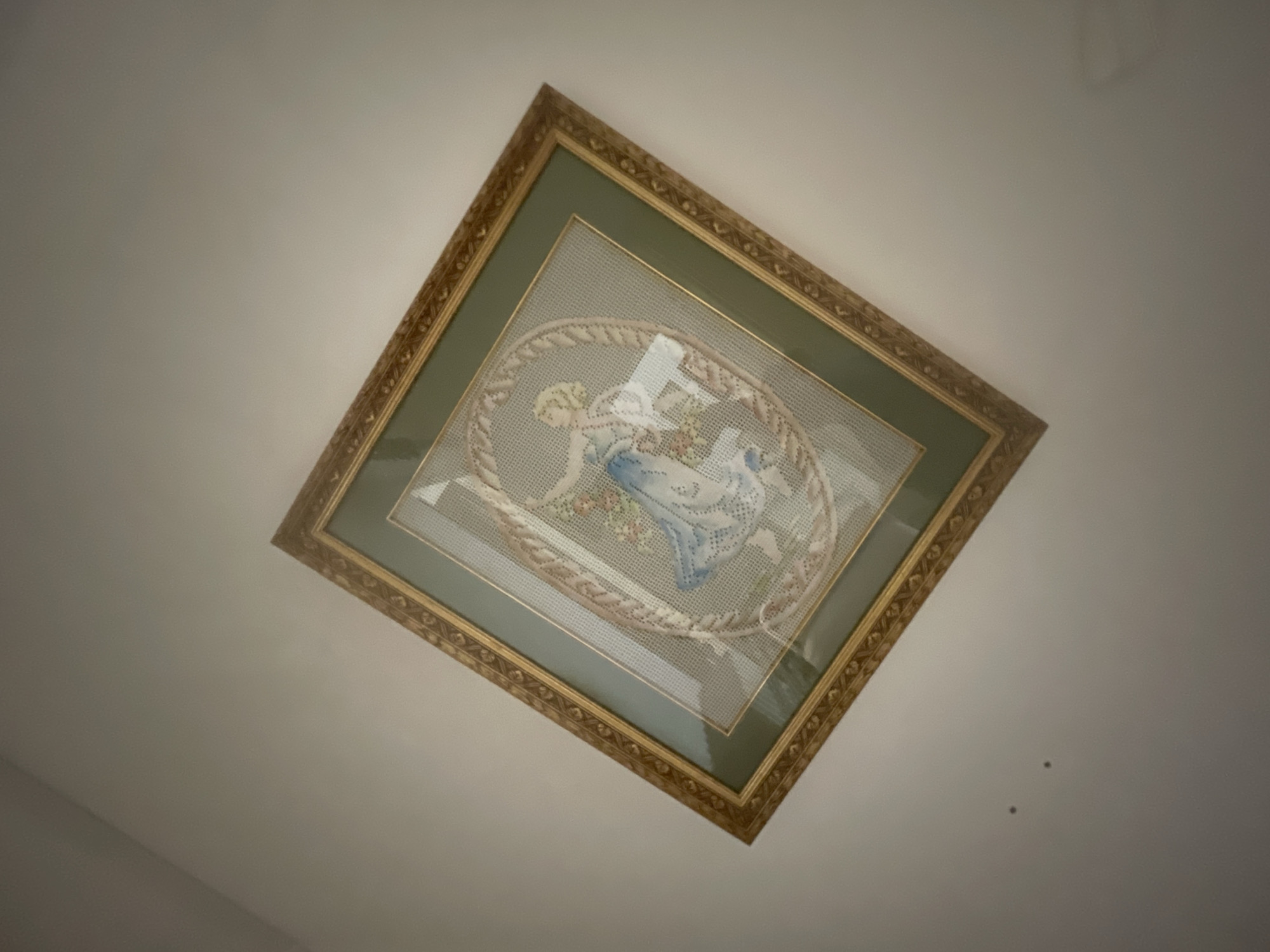 A framed needlepoint artwork hangs on a plain, light-colored wall. The artwork depicts a woman in a blue and white dress, possibly dancing, with a floral motif around her. The frame is ornate and gold-colored, with a darker green matting surrounding the needlepoint. The image is taken from a low angle, with the artwork tilted, and the wall is illuminated by a light source that creates a soft gradient. Two small dark spots are visible on the wall below the artwork. The overall scene suggests an indoor setting, likely a room in a home or gallery.