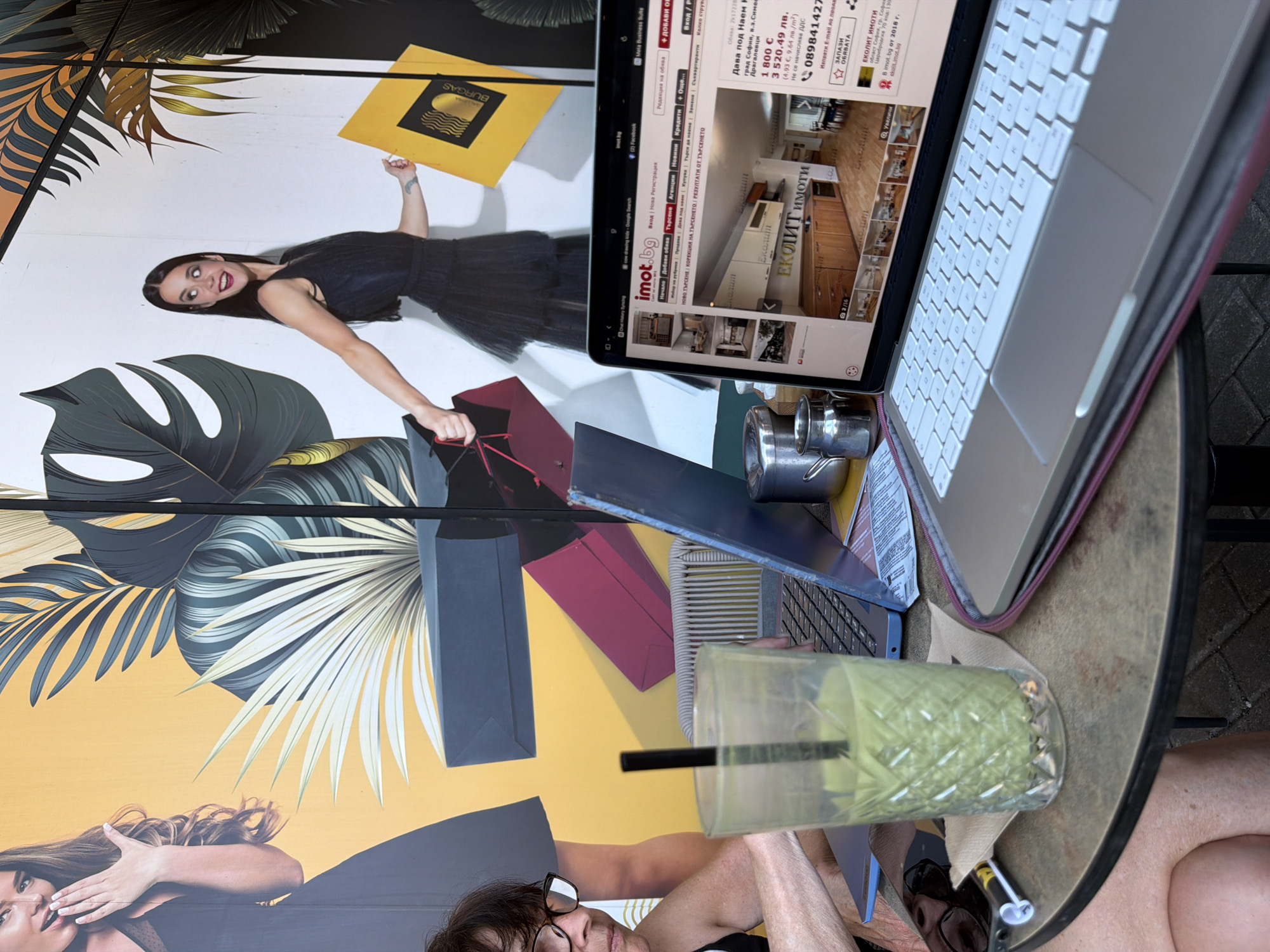 The image shows a close-up of a laptop screen displaying a real estate listing on the website imot.bg. The laptop is open and placed on a table next to a glass of green beverage with a straw and a person's lap and arm. The background appears to be a mural with images of women and tropical plants. The laptop screen shows details of an apartment for sale in Sofia, Bulgaria, including images of the interior, price, and contact information. The time and date on the laptop screen appear to be December 2, 2018, at 09:22.