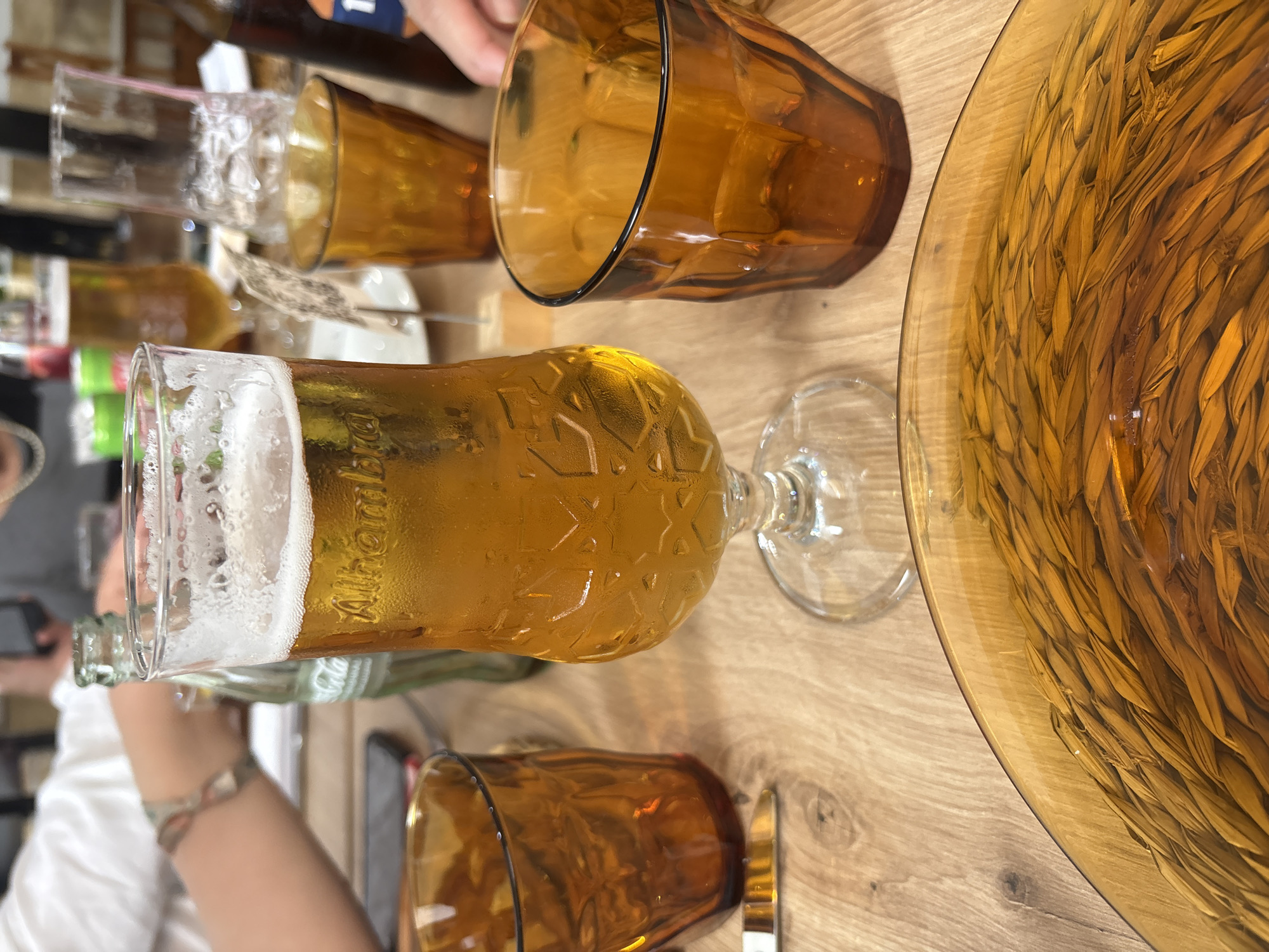 A wooden table displays glasses and bottles, including a beer glass with the "Alhambra Especial" logo filled with a golden liquid and a thick white foam head, along with three amber-colored tumblers, a clear drinking glass, a green beer bottle, and a large amber-colored plate with a woven straw design beneath a transparent layer. The edge of a person's arm with a bracelet is visible in the lower left corner, and another person's arm is partially visible in the upper center. The location is likely a bar or restaurant in Córdoba, Spain, given the Alhambra beer, popular in the region.