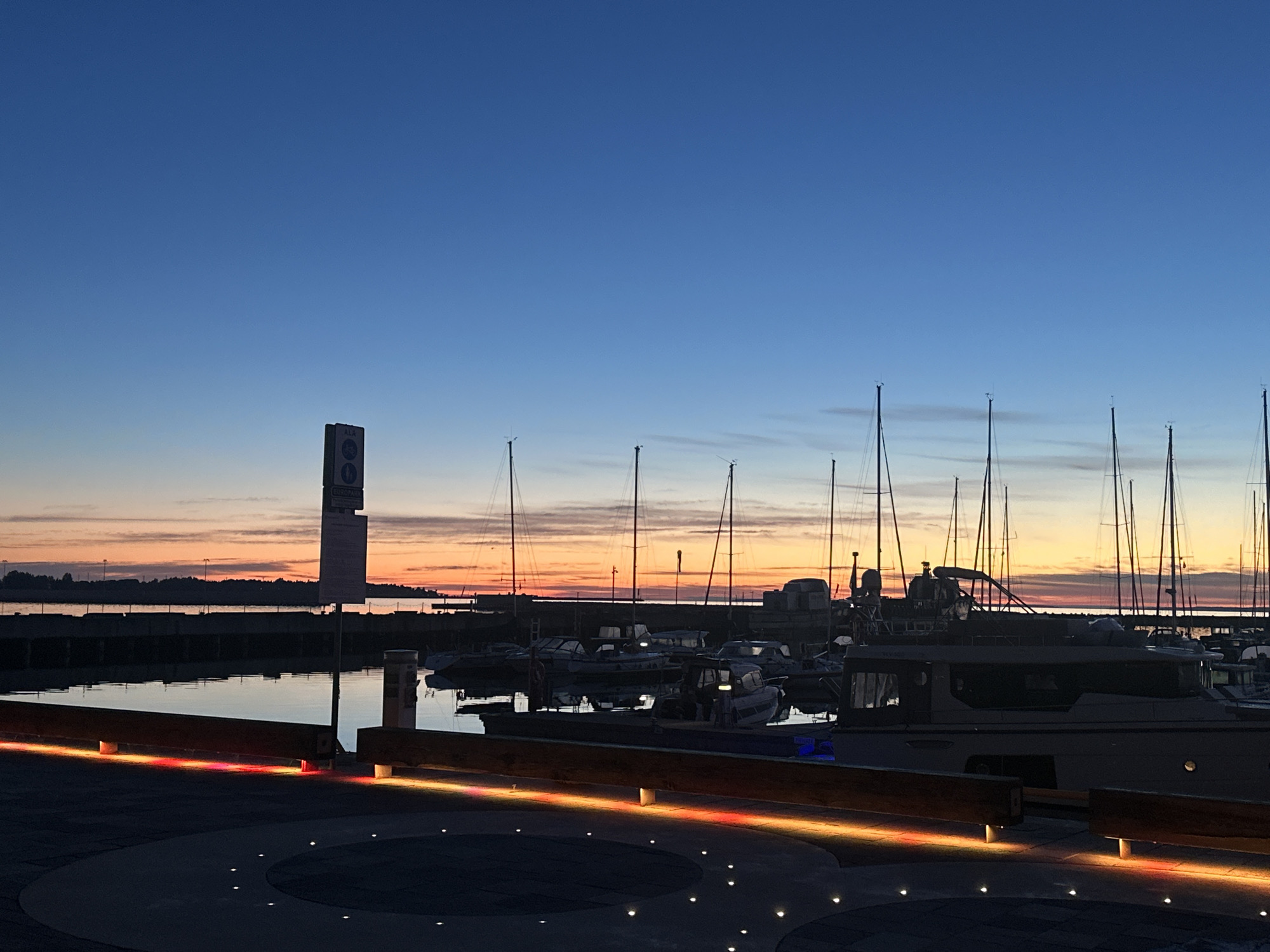 This image captures a serene twilight scene at a marina, likely in Tallinn, Estonia, given the presence of a sign that appears to be in Estonian. The sky transitions from a deep blue at the top to soft hues of orange and pink near the horizon, indicating either sunrise or sunset. A calm body of water, reflecting the sky and the marina's lights, fills the middle ground.
Dozens of sailboats and motorboats are docked in the marina, their masts forming a delicate silhouette against the colorful sky. Some of these boats appear to be illuminated from within. In the foreground, a paved area with embedded circular lighting features is visible. A wooden structure, possibly a bench or railing, runs along the edge of the paved area, illuminated by a strip of red and yellow lights. A signpost with multiple signs stands to the left of the frame, one of which seems to be a traffic or directional sign. The overall atmosphere is peaceful and tranquil, characteristic of a quiet evening in a harbor. No people are visible in the scene.