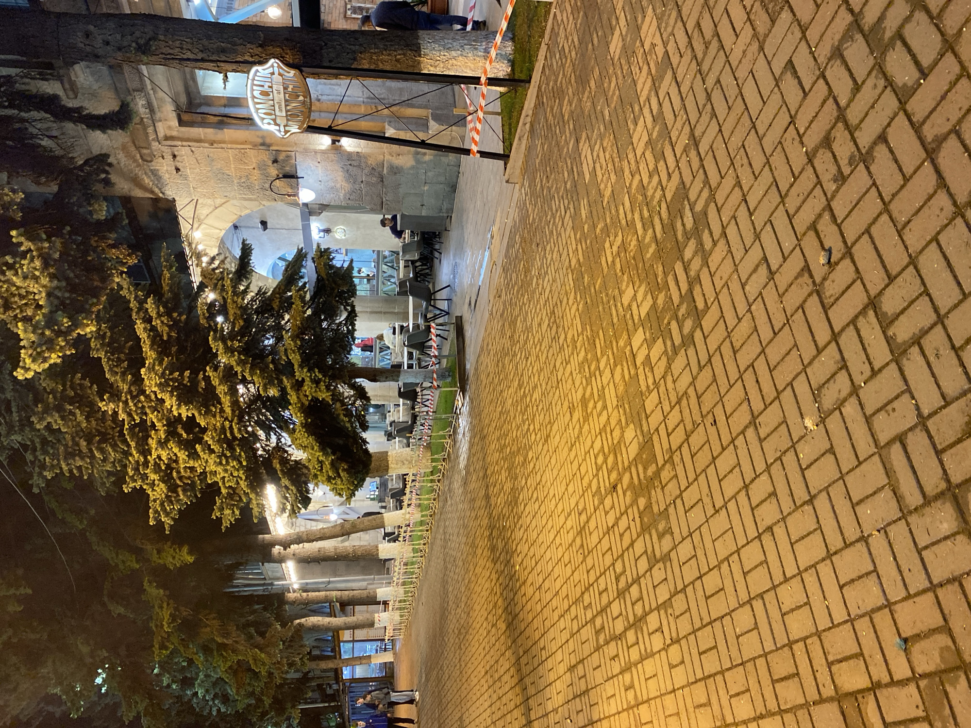 The image depicts an outdoor seating area of a restaurant at night in Gyumri, Armenia. The foreground is a paved sidewalk with interlocking rectangular bricks, wet and reflecting the warm glow of overhead lights and streetlights. Beyond the sidewalk, there is a row of tables and chairs set up for outdoor dining, some of which are occupied by patrons. A line of trees with illuminated foliage separates the sidewalk from the restaurant's outdoor seating area. The restaurant itself appears to be a building with arched windows and stone architecture. A prominent sign with the word "PONCHIK" is visible, indicating the name of the establishment. A person is walking on the sidewalk in the background, and another individual is standing near the entrance of the restaurant. Red and white tape is used as a barrier in some areas. The overall atmosphere is one of a relaxed evening at a dining establishment.
