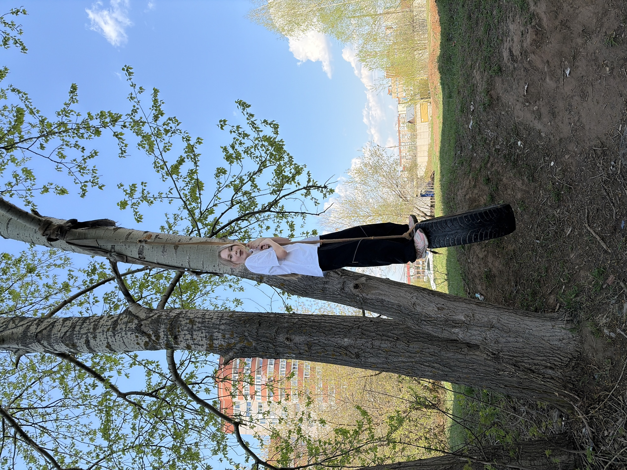 A young woman is suspended on a tire swing between two tree branches in an outdoor location. The tire is hanging on a rope. The woman wears a white t-shirt, black pants, and pink sneakers. The trees are large and mature, with green leaves sprouting. In the background, there is an apartment building with a red roof, partially obscured by foliage. The location is Izhevsk, Russia. The ground is covered in dirt and vegetation. The sky is blue with some clouds.