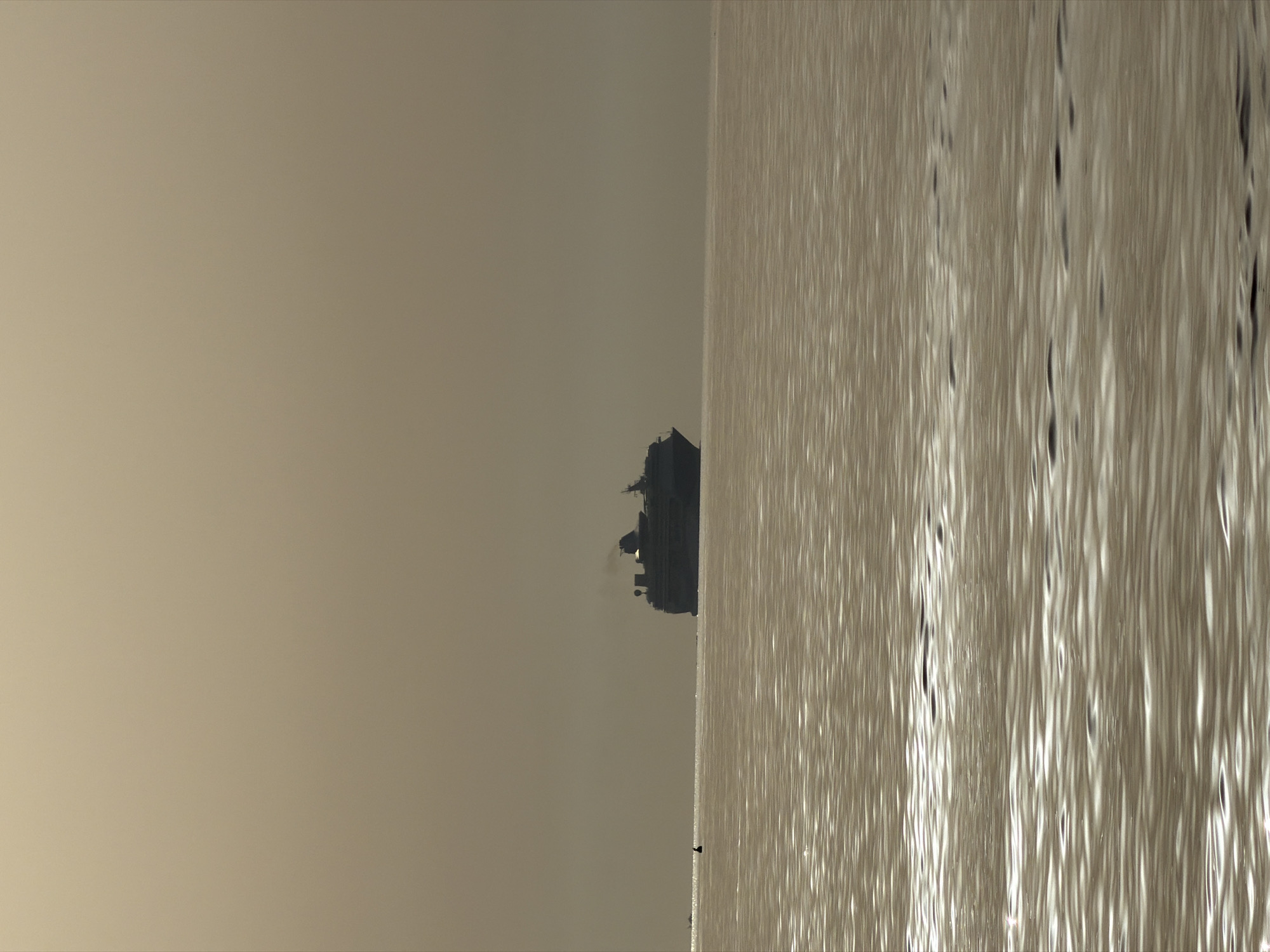 This image depicts a cruise ship silhouetted against a hazy, sepia-toned sky over a calm body of water. The ship, appearing dark and indistinct, is positioned towards the right side of the frame. Its silhouette suggests multiple decks and at least one smokestack emitting a plume of smoke. In the foreground, the water exhibits a textured surface with ripples that catch the light, creating a shimmering effect. A small, dark, buoy-like object is visible in the water to the left of the ship, closer to the horizon. The overall atmosphere is muted and serene, with the soft light and lack of sharp details suggesting either dawn or dusk, or a day with overcast or foggy conditions. The scene is devoid of human figures. This could be a view from the coast or a boat in the Gulf of Olbia, Sardinia, Italy, where such large vessels are common.