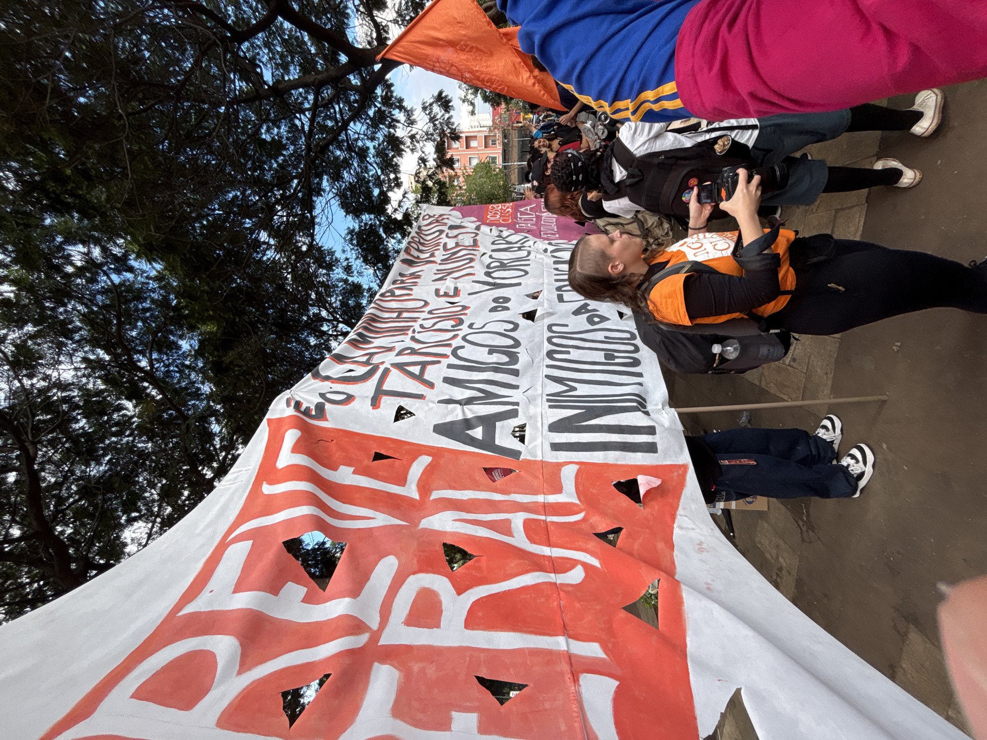 A low-angle, outdoor shot depicts a political demonstration in what appears to be the city of São Paulo, Brazil. A large white banner with cut-out triangular shapes is stretched out, displaying painted text in Portuguese, including the words "AMIGOS" (friends) and "INIMIGOS" (enemies). The banner is held by several people. A woman in an orange shirt takes a picture with a camera. Other individuals are milling around. Trees and buildings are visible in the background.