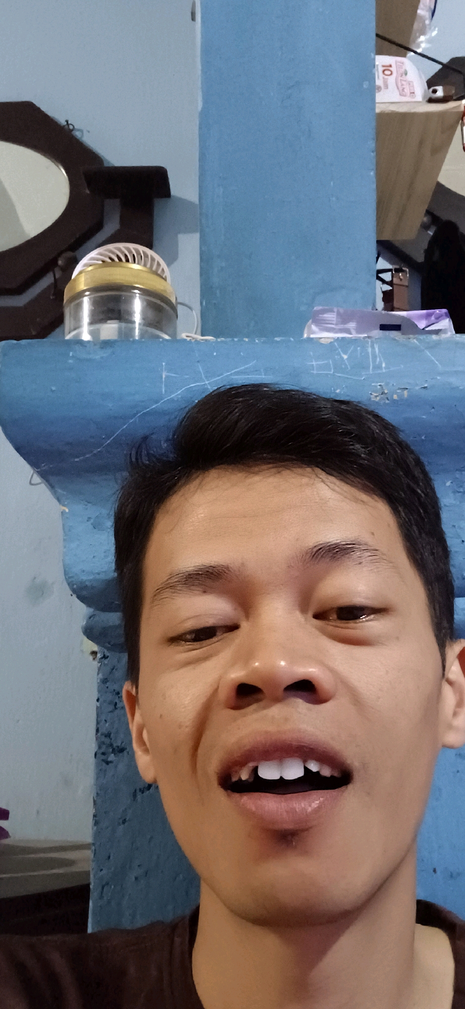 A medium shot photograph shows a young man looking directly at the camera. The man has short, dark hair and is looking slightly to his right. His mouth is open, revealing white teeth. The background of the image is a blue wall, and a shelf is visible above the man's head. On the shelf are several objects, including a fan and a packet of tissues. The photograph appears to have been taken indoors.