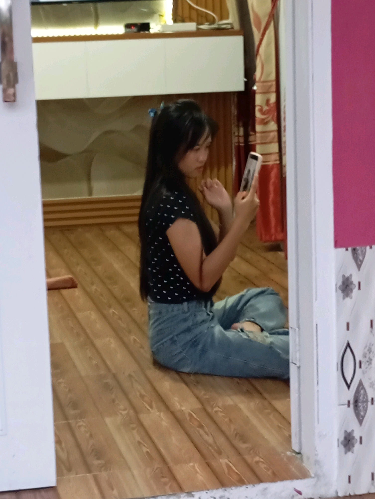 A young woman is sitting on a wooden floor, holding a smartphone in her right hand and looking at its screen. She is wearing a black polka dot shirt and ripped blue jeans. The room appears to be indoors, with wooden flooring and a white wall to the left. There are curtains and a cabinet in the background. The image appears to be taken in Muncang, Indonesia.