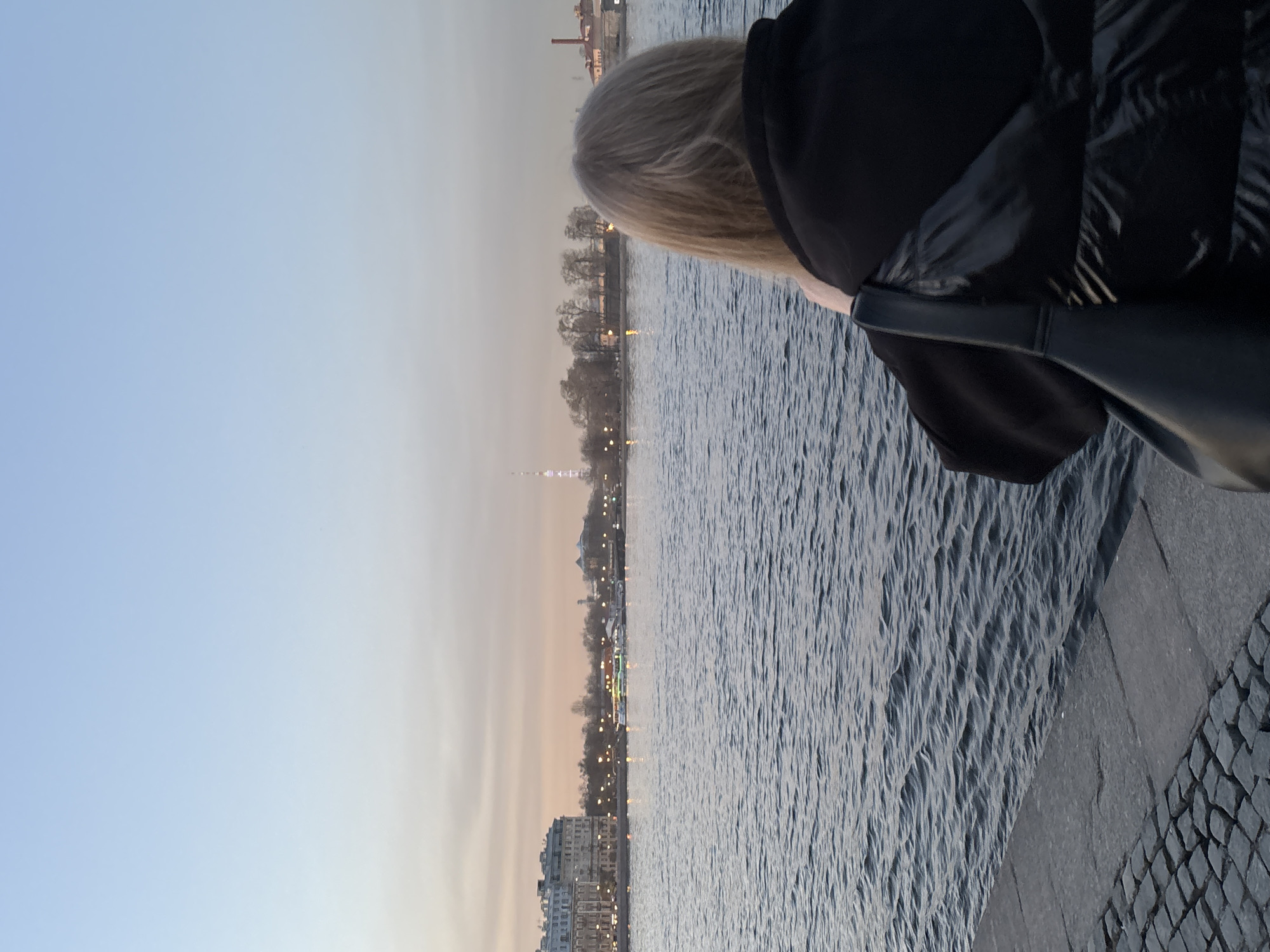 A person with blonde hair, wearing a black jacket and purse, stands on a grey stone embankment overlooking a body of water with choppy waves. Across the water is a cityscape featuring buildings and a tall, thin tower that is identified as the Saint Petersburg TV Tower. The time of day appears to be dusk, as evidenced by the soft, muted light in the sky. The sky is a light blue color that fades to a warm hue near the horizon.