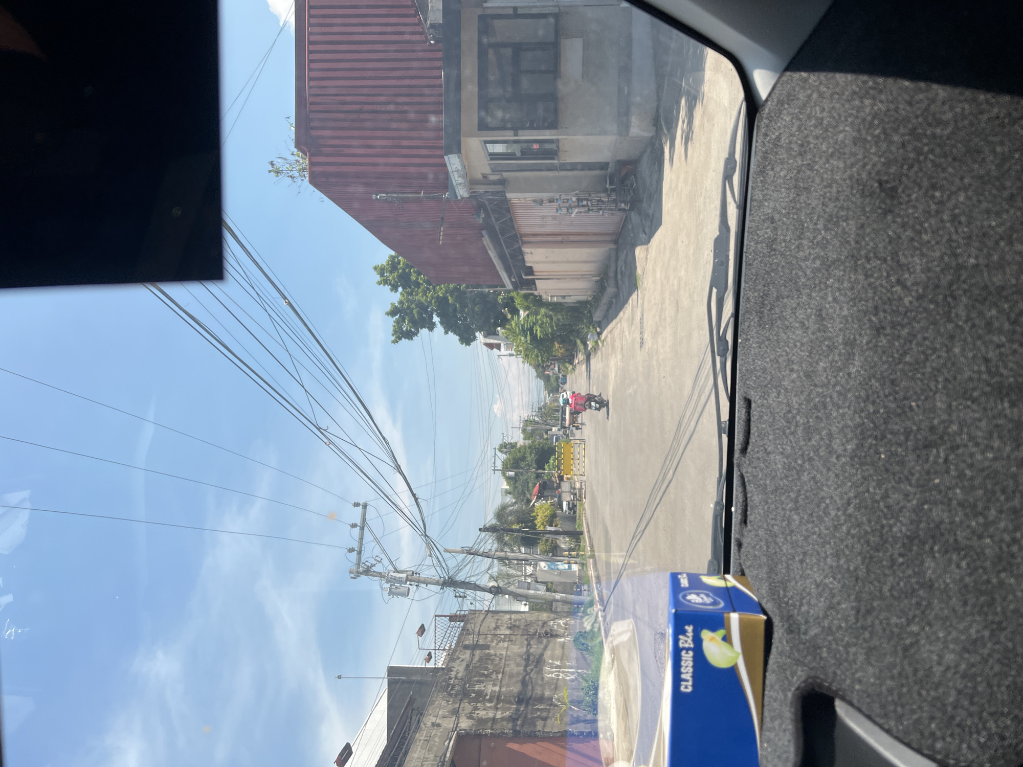 The image presents a daytime street view in Davao City, Philippines, captured from inside a vehicle. The sky is bright blue with scattered light clouds, indicating sunny weather.
In the immediate foreground, a dark gray, textured dashboard is visible, topped by a blue "CLASSIC Blue" tissue box featuring a green lime graphic. The vehicle's windshield wipers are visible at the base of the windshield.
The street, appearing to be concrete or asphalt, extends into the mid-distance. On the left side, a tall, weathered concrete wall with dark stains and markings runs alongside the road. Numerous utility poles, constructed of both wood and concrete, line this side, supporting a dense array of overhead power and communication lines; one pole features a transformer.
On the right side of the street, a large building with a reddish-brown, vertically corrugated metal facade is prominent. Adjacent to it is a smaller, light-colored concrete structure, possibly a guardhouse, with dark-tinted windows. A light-colored concrete fence with a brown gate is visible further down this side, with some metal racks or shelving positioned outside the gate.
Towards the middle ground, a yellow barrier or barricade partially spans the street. A lone individual on a red motorcycle, wearing a pink or red shirt, is observed driving away from the camera toward this barrier. Another small vehicle, consistent with a tricycle, is partially visible near the barrier on the right. No other individuals or specific interactions are discernible.