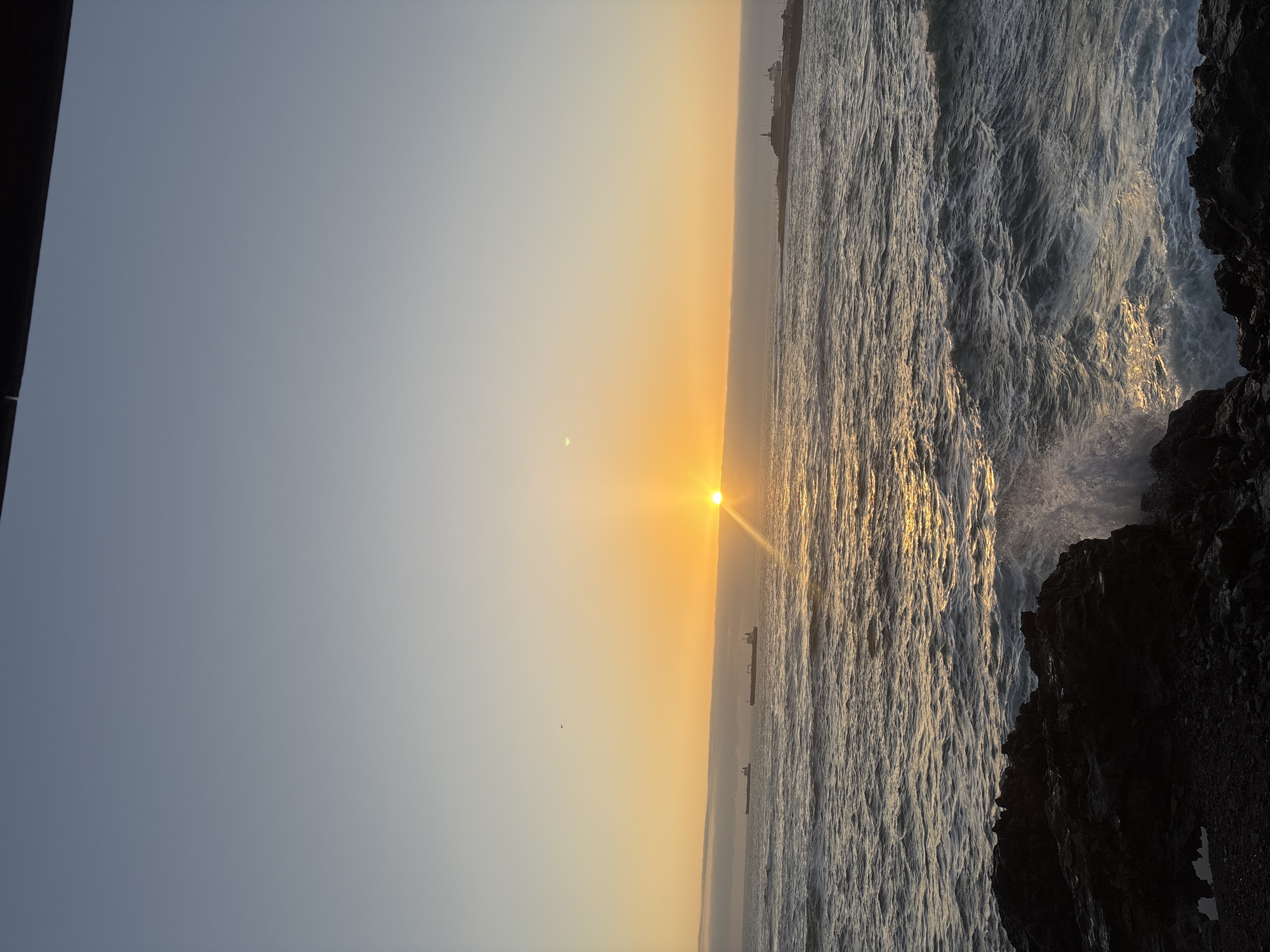 The image captures a sunset scene in Arica, Chile, featuring the Pacific Ocean. The sun is positioned near the horizon, casting a golden glow across the water. Waves are breaking against a rocky coastline in the foreground. Two cargo ships can be seen in the distance. A crescent moon is faintly visible in the sky. The lighting suggests a dusk or dawn setting. The angle of the photo shows the photographer was standing on the cliff above the rocky coastline.