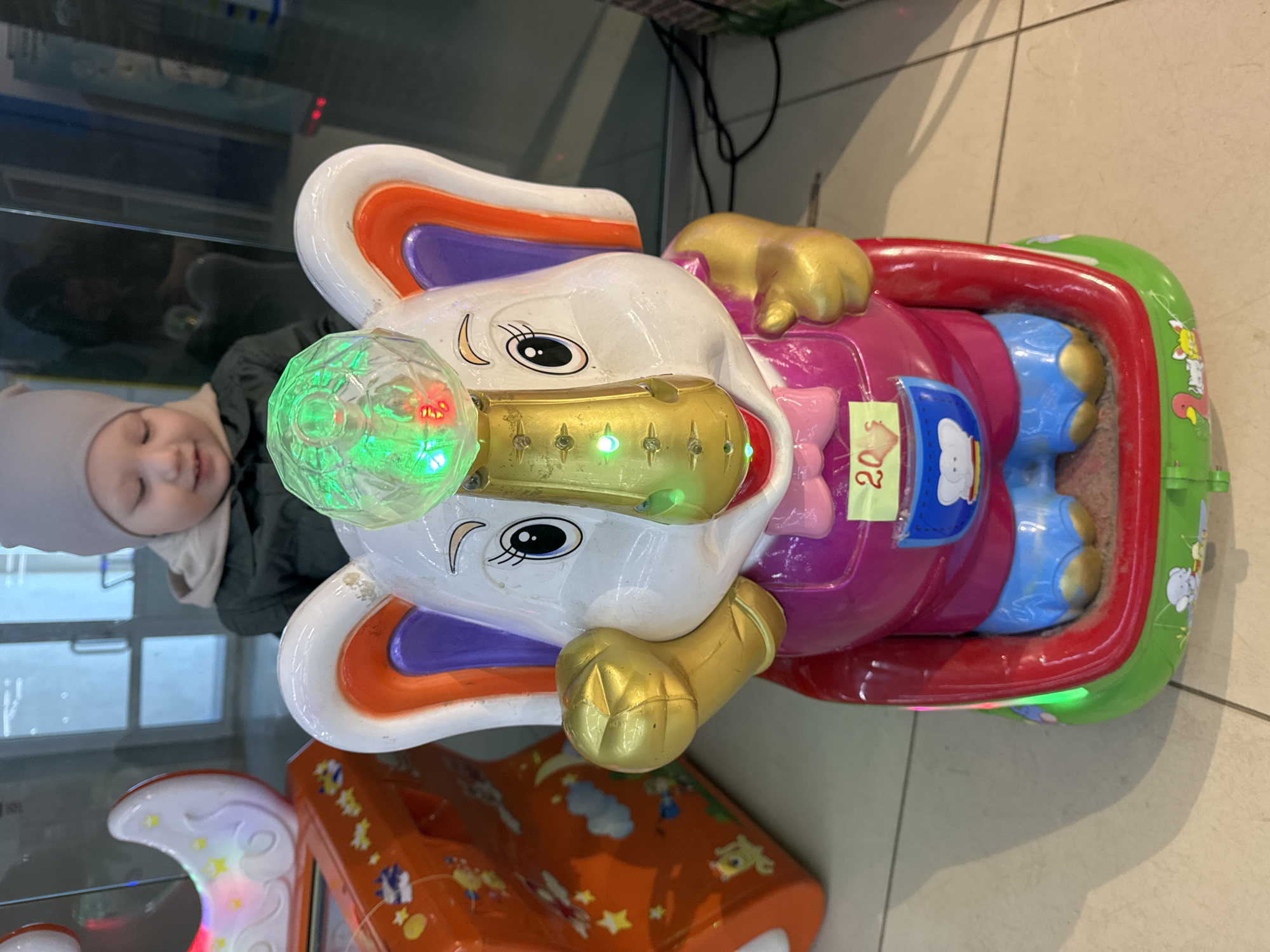 The image shows a young child positioned next to a brightly colored, coin-operated elephant ride in what appears to be an indoor public space, possibly a shopping mall or entertainment center in Chaikovsky, Russia. The child, wearing a light grey beanie and a dark jacket, is visible on the left, looking towards the ride with a joyful, smiling expression.
The prominent elephant ride is predominantly white, featuring large ears with orange and purple accents. Its trunk and feet are painted a metallic gold. The elephant's body, designed as a seat, is pink with blue "pants" for its legs, encased by a red safety bar and a vibrant green base decorated with cartoon animals. A disco ball-like light fixture on the elephant's head emits green and red light, complemented by smaller blue lights along its trunk. A yellow sticker on the side of the ride clearly displays "20₽," indicating the price in Russian Rubles.
Part of another orange-colored ride, decorated with stars and clouds, is visible in the bottom left corner, suggesting a collection of such amusements. The floor is tiled, and the background shows reflections, consistent with an indoor setting. The scene captures a moment of childhood amusement and joy.