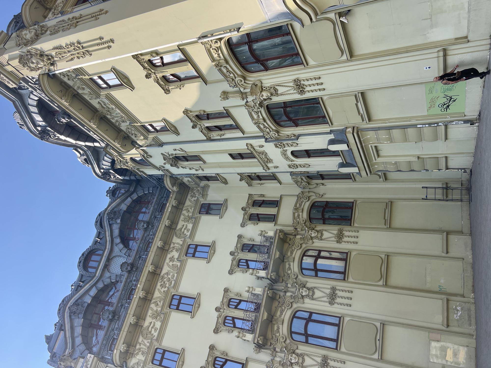 The image shows the exterior of a light yellow building with ornate architectural details in Odesa, Ukraine. The building has a curved, mansard roof with decorative elements and multiple windows. The facade is adorned with reliefs of faces, foliage, and other sculptural elements. The lower part of the building features large, arched windows and a few flat, recessed panels. A person with tattoos and a shaved head is standing near the right edge of the building, leaning against the wall. They are wearing a black tank top, black pants, and a brown fanny pack. Graffiti is visible on a green section of the wall near the person. A metal ladder is propped up against the building on the left side. The sky above is clear and blue.