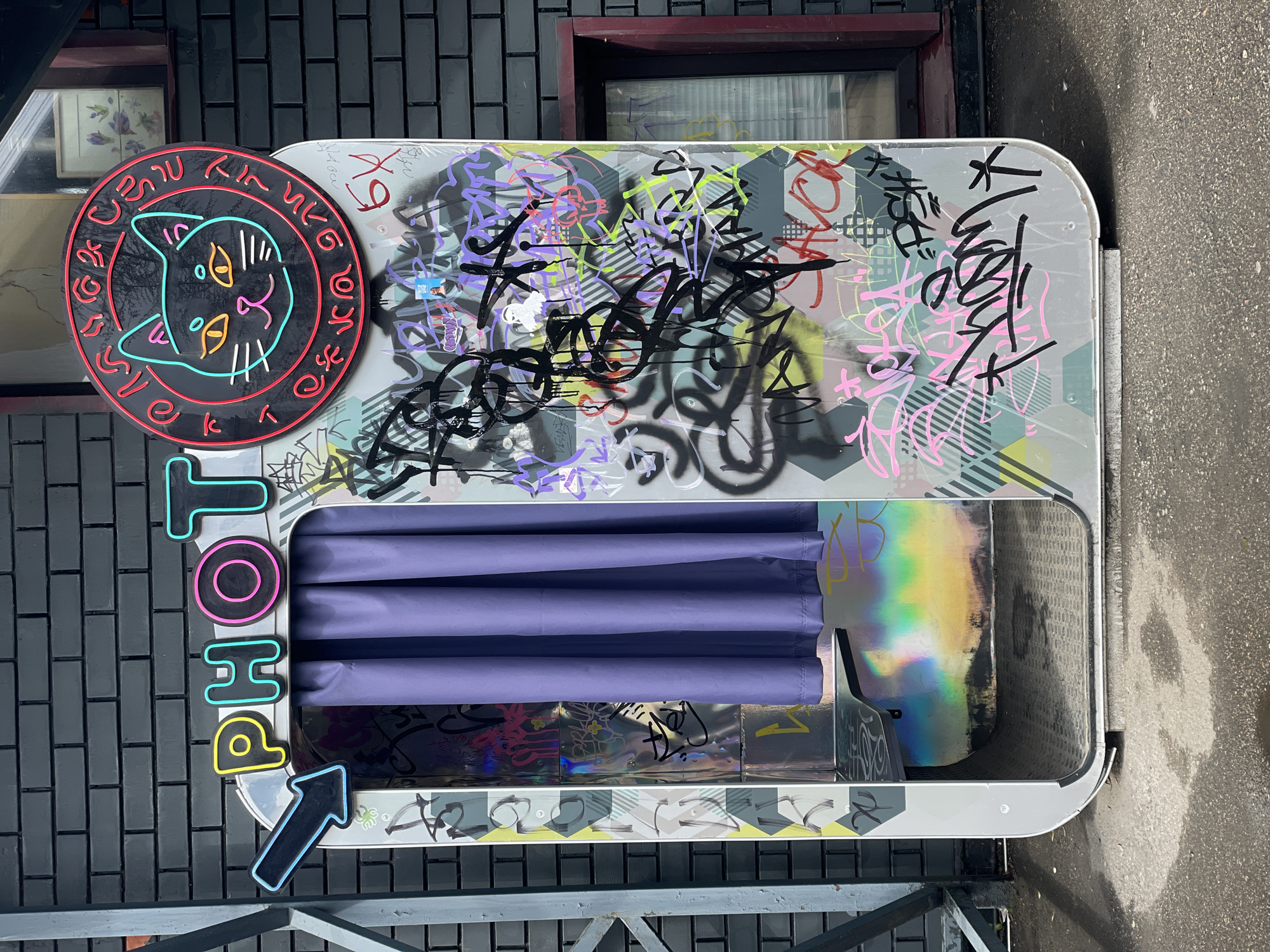 A photobooth stands against a dark brick building, adorned with graffiti and stickers. The booth's interior is concealed by purple curtains. To its left, a neon sign spells "PHOTO" vertically with an arrow pointing towards the booth. Above the booth, a round neon sign depicts a cat's face within a circle of unidentifiable writing. The surrounding environment is an urban setting, with wet pavement visible to the right and part of a metal fence in the foreground. While the location is identified as Khimki, Russia, this is not evident from the visual elements alone.