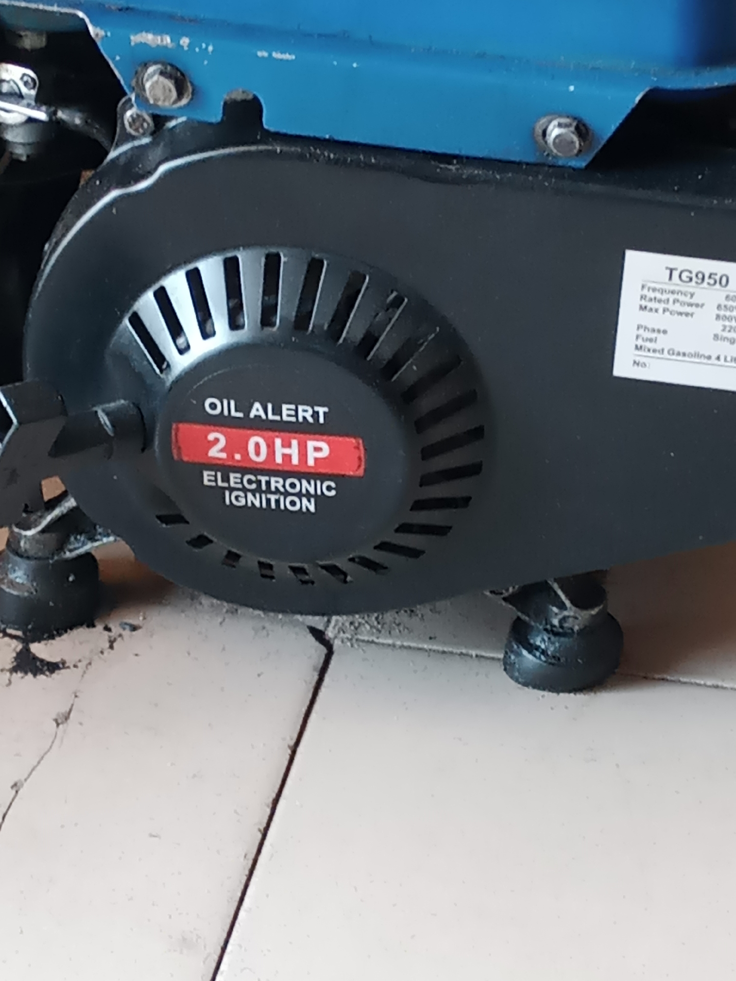 This is a close-up, eye-level shot of a blue and black portable generator, likely located on a tiled floor in Jalingo, Nigeria. The generator's black engine housing is prominent, featuring a circular fan cover with metal slats. A red sticker is affixed to the fan cover, displaying "OIL ALERT" in white text above "2.0 HP" in larger white text. Below this, "ELECTRONIC IGNITION" is also in white lettering. To the right of the fan cover, a white label with black text lists specifications for a "TG950" model generator, including "Frequency 60Hz", "Rated Power 650W", "Max Power 800W", "Phase Single", "Fuel Mixed Gasoline 4 Liters", and "No:". The generator is supported by four black rubber feet, one of which is clearly visible at the bottom right. The background consists of light-colored tiles, some of which appear cracked, suggesting an indoor or covered outdoor setting. The overall condition of the generator appears used, with some dust and grime visible on the engine housing and feet.