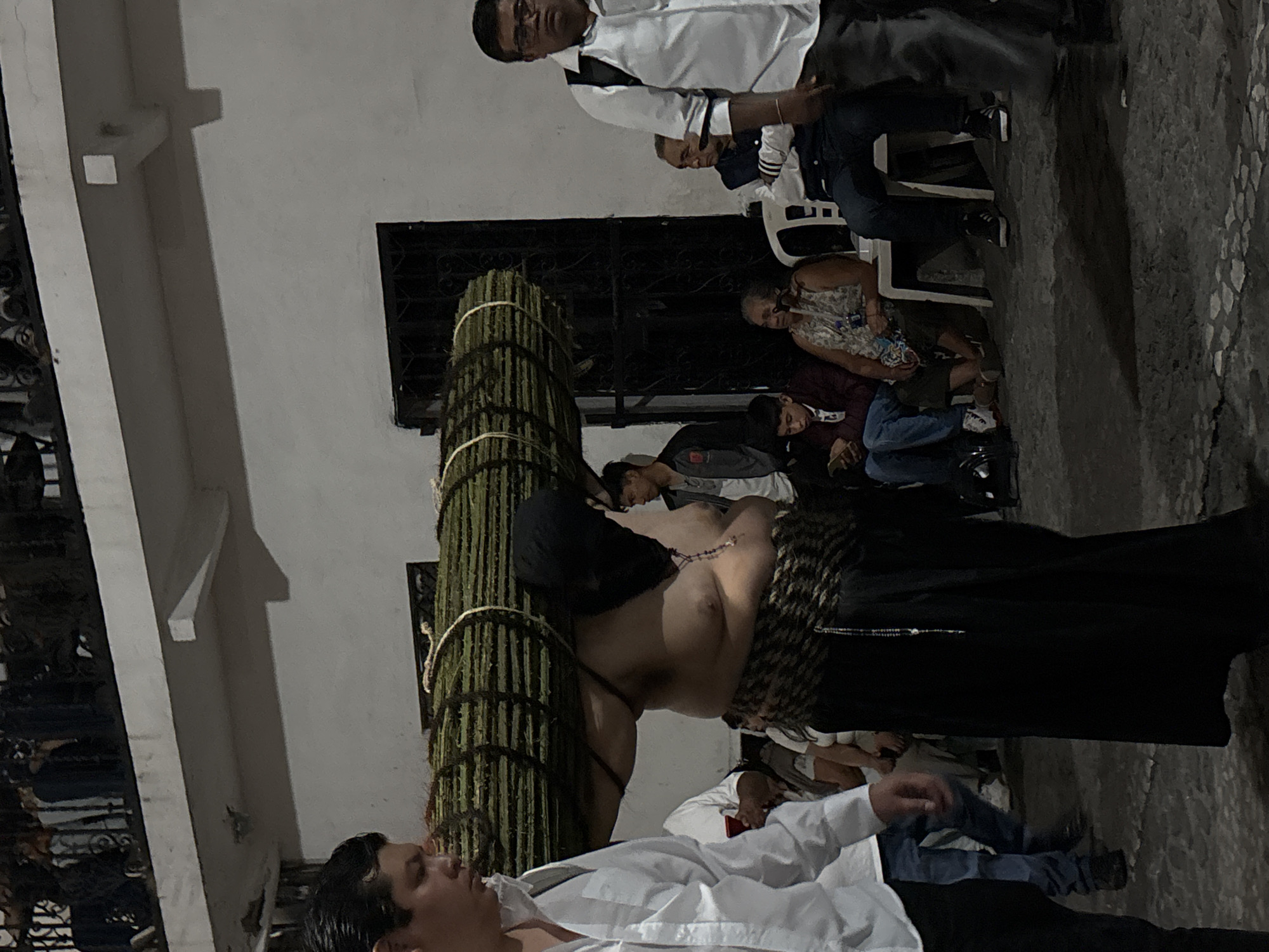 In Taxco, Mexico, a man, partially unclothed and carrying a large bundled stick cross, stands near a wall. People sit and stand nearby, observing. The setting appears to be outdoors at night.