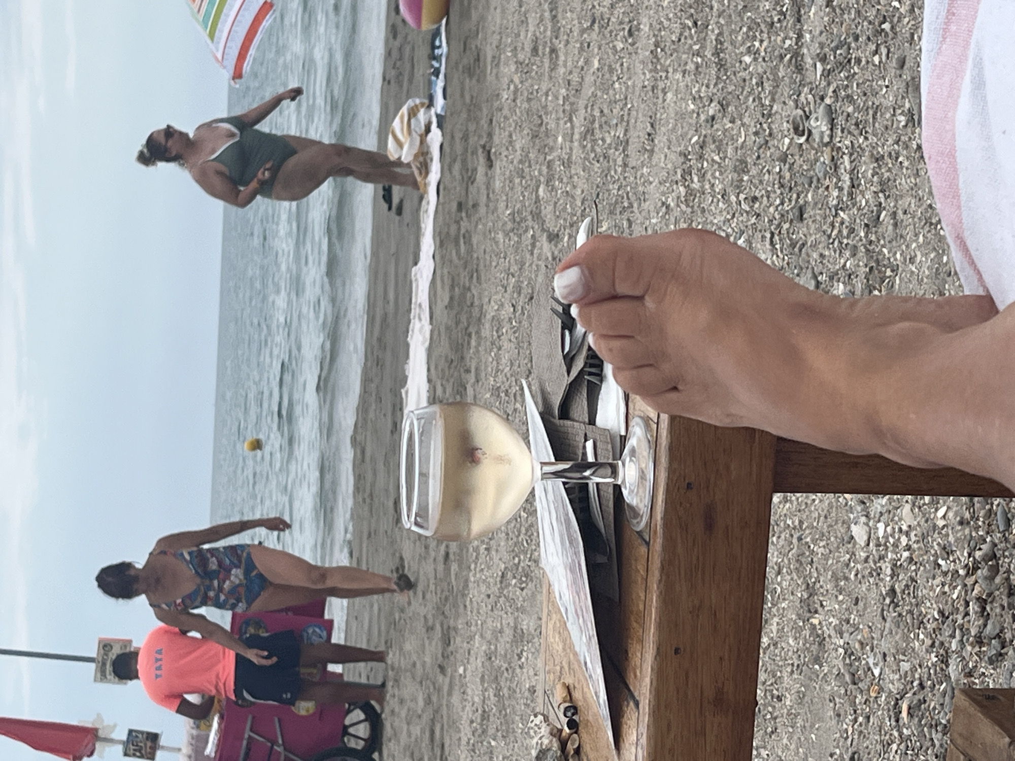 The image depicts a beach scene in Frontignan, France, with the Mediterranean Sea in the background. In the foreground, a person's bare foot rests on a towel, partially obscuring a wooden table. On the table, a glass of white wine and some discarded cigarette butts are visible. Further back on the sand, two women are walking towards the sea. One woman wears a patterned swimsuit, and the other wears a green swimsuit. To the left, a person in an orange t-shirt stands near a pink ice cream cart. The sky is overcast, and a yellow buoy floats in the water. An umbrella with colorful stripes is partially visible on the right side of the image.