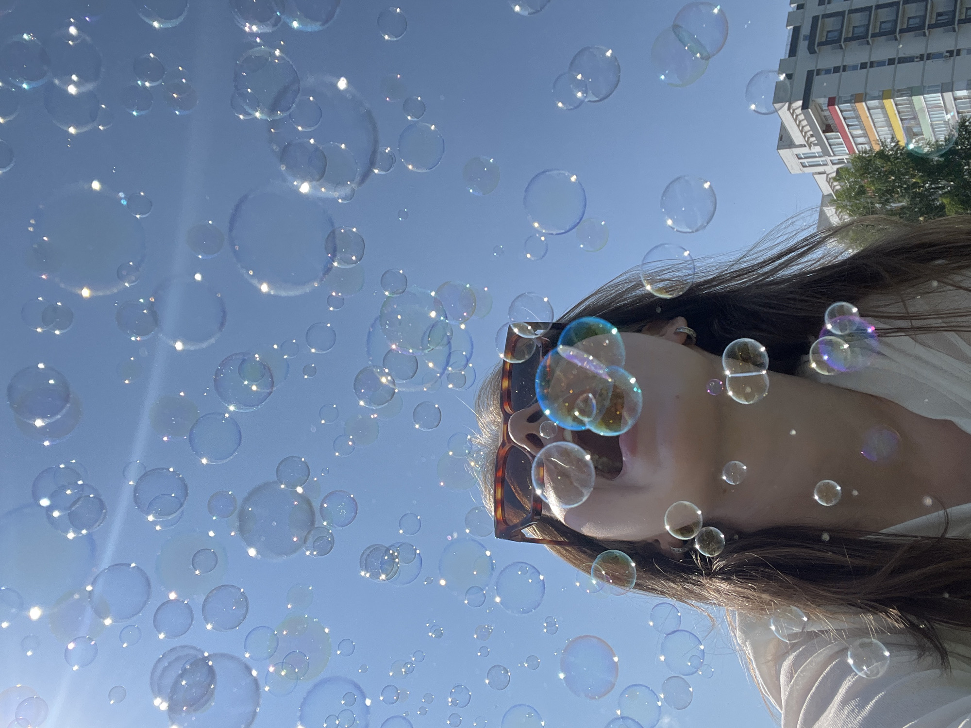 The image captures a young person blowing bubbles against a bright blue sky. The person is wearing sunglasses with tortoise-shell frames and a light-colored shirt. Bubbles of various sizes are scattered throughout the frame, catching the sunlight and reflecting iridescent colors. In the background, a modern apartment building with colorful accents and lush green trees can be seen, suggesting an urban or suburban setting. The low-angle shot emphasizes the vastness of the sky and the playful atmosphere of the scene. This image appears to be taken in Chaikovsky, Russia, as the architecture and environment are consistent with the city's urban landscape.
