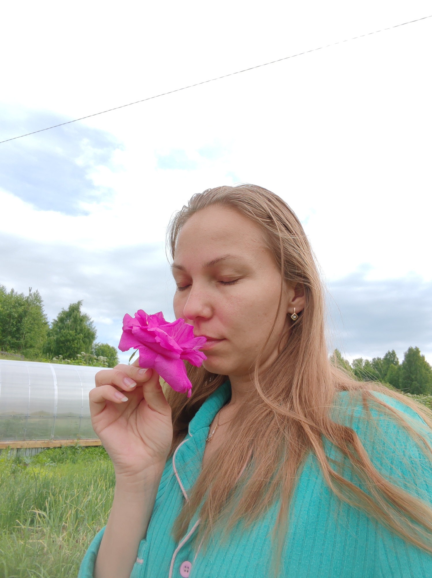 A medium shot shows a fair-skinned woman with long, blonde hair, wearing a turquoise pajama top with pink trim and buttons, holding a vibrant pink rose to her nose and smelling it with her eyes closed. She is wearing a small, diamond-shaped gold earring in her right ear. She is outdoors, with a blurry background that includes a greenhouse structure, tall green grass, trees, and a cloudy sky with a wire running across it. The scene appears to be a backyard or garden setting in or near the city of Chukhlem, Russia.