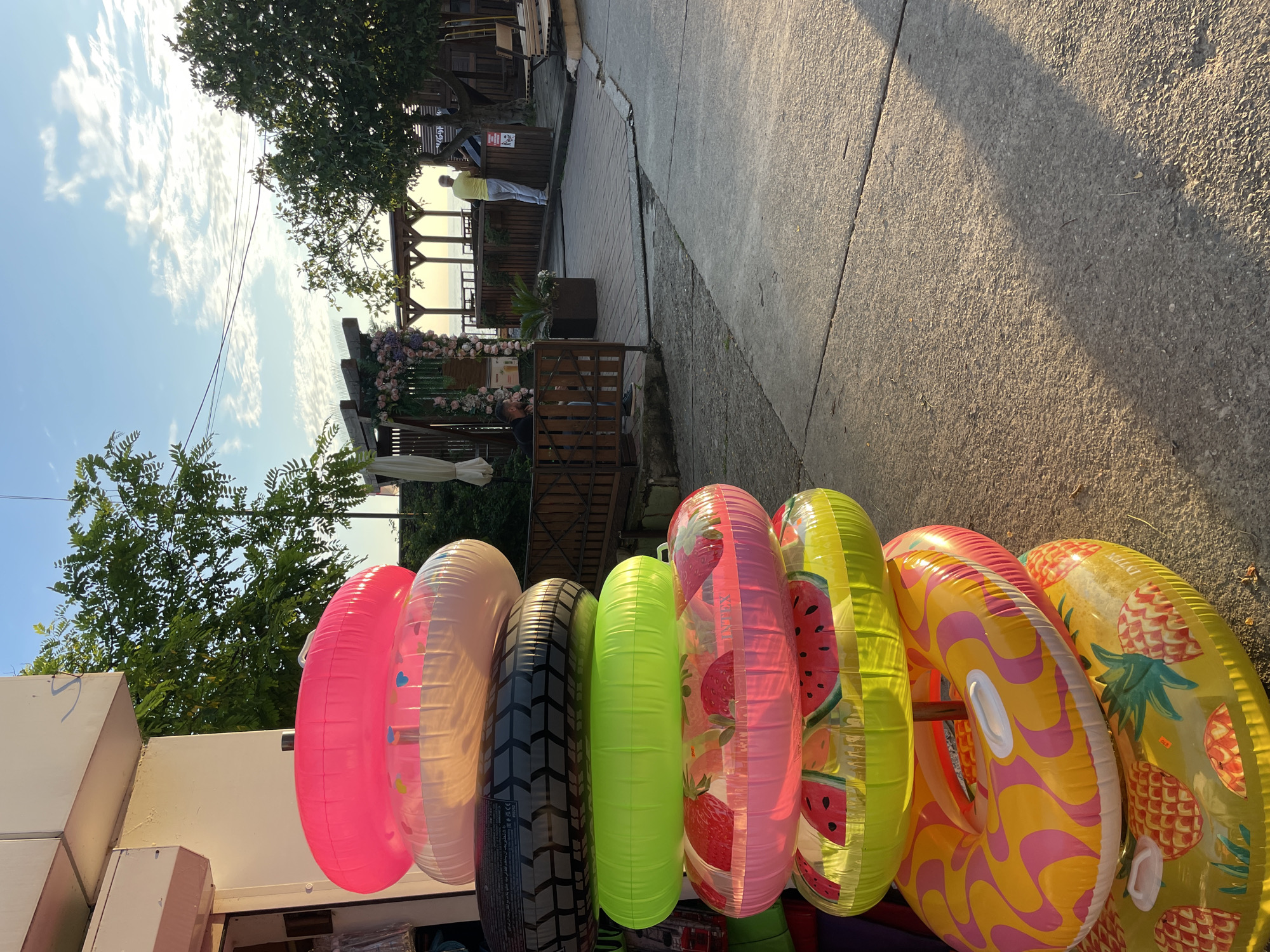 A stack of brightly colored inflatable swimming rings is positioned in front of a building with white siding. The rings are decorated with various patterns, including a tire tread, strawberries, watermelons, and pineapples. Behind the rings, a pathway leads towards a wooden deck with tables and chairs, suggesting an outdoor seating area. In the background, trees and a clear sky with scattered clouds indicate a sunny day, likely in a resort area given the presence of swimming rings. The scene appears to be in Sochi, Russia, as the context suggests a location associated with tourism and outdoor recreation. Several people are visible in the background, including one person in a yellow shirt standing on the deck and another in a black shirt sitting near a wooden fence.