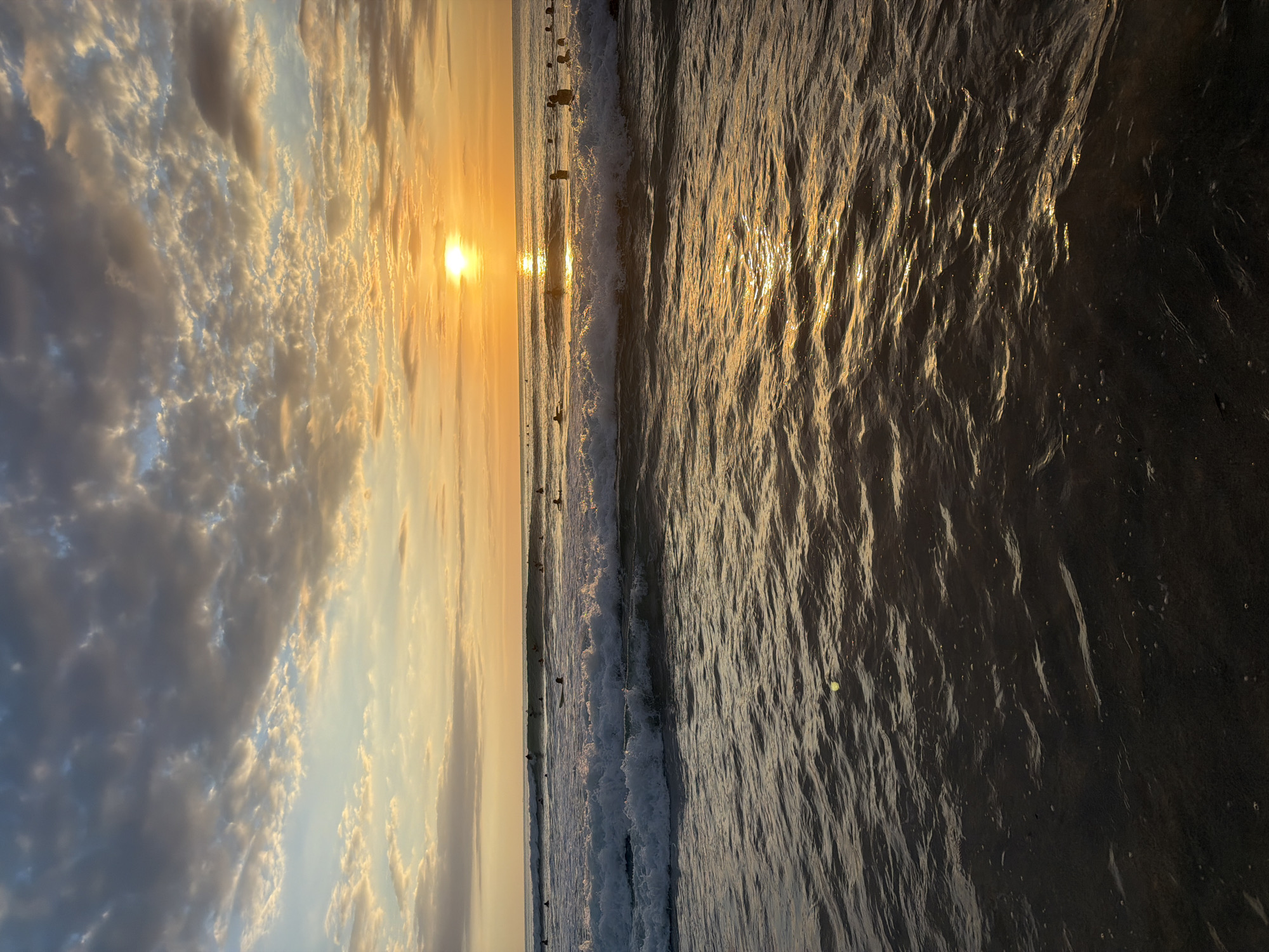 The image shows a beach scene in Kuta Selatan, Indonesia, at sunset. The sky is filled with clouds, illuminated in hues of gold and orange by the setting sun. Below the sky, the ocean reflects the golden light, with waves breaking along the shore. Numerous people, possibly surfers, are visible in the water near the shoreline.