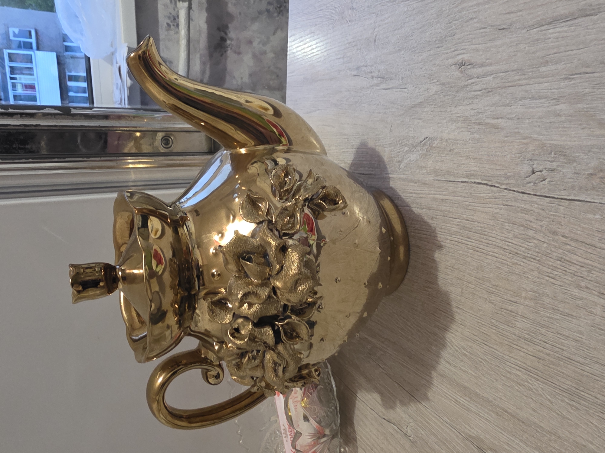 A metallic gold teapot with embossed floral designs sits on a light-colored wooden surface, which appears to be a table. The teapot has a curved handle, a prominent spout, and a lid topped with a small decorative element. In the background, a window reveals a glimpse of a building with balconies and windows, suggesting an urban setting. The overall scene is an indoor still life, possibly a domestic setting.