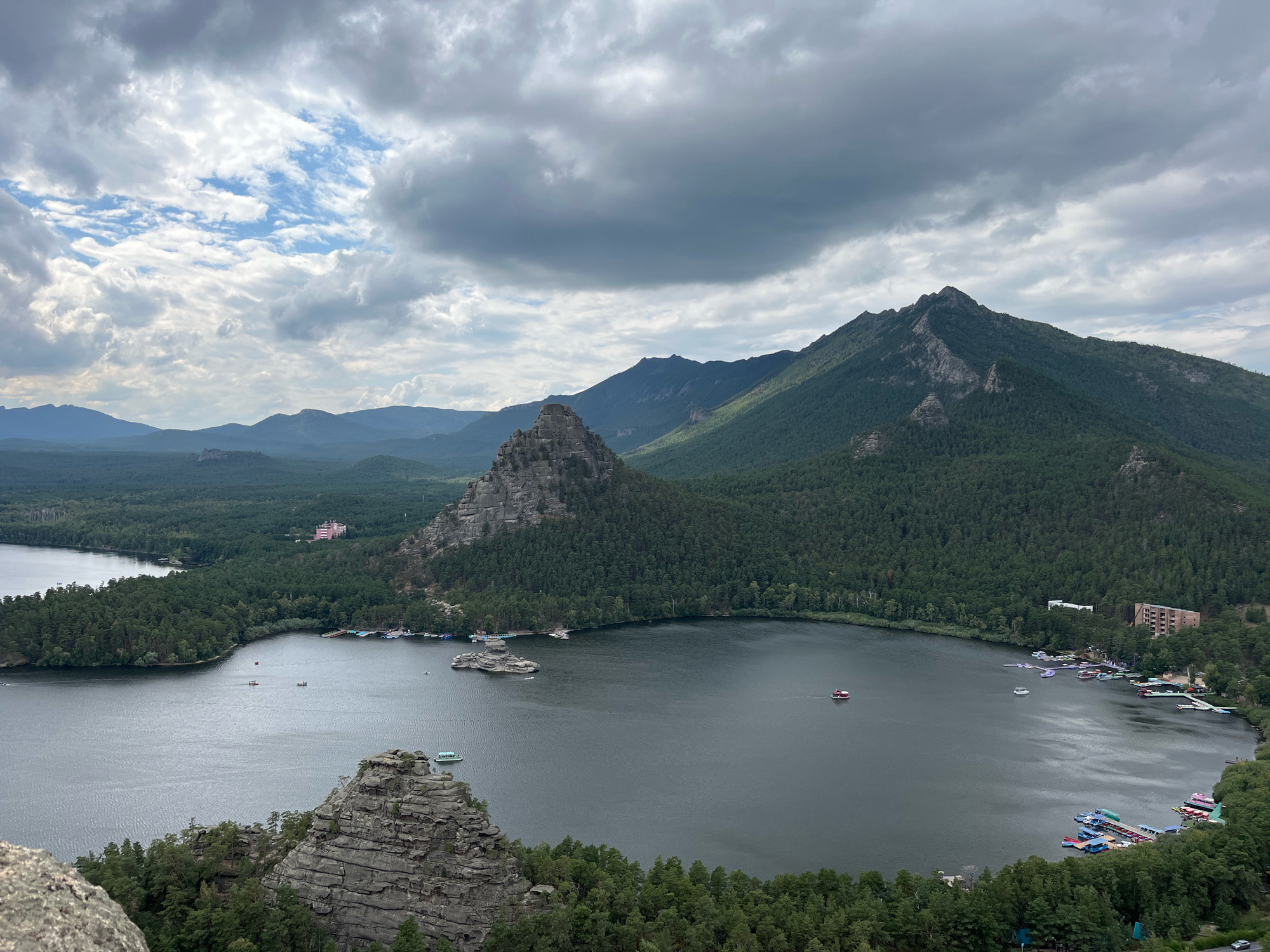 This is an aerial view of a lake in a mountainous, forested region. The lake is dark and calm, with several small boats and a larger red boat visible on its surface. The surrounding landscape is covered in dense green forest, with rugged, rocky mountains rising in the background. The sky is overcast with dramatic clouds. On the far shore of the lake, there are several buildings, including a pink hotel-like structure and some resort-style accommodations with docks. The foreground features a rocky outcrop with trees. The location is identified as Burabay National Park, located within the Зелёный Бор ауылдық округі, Kazakhstan.