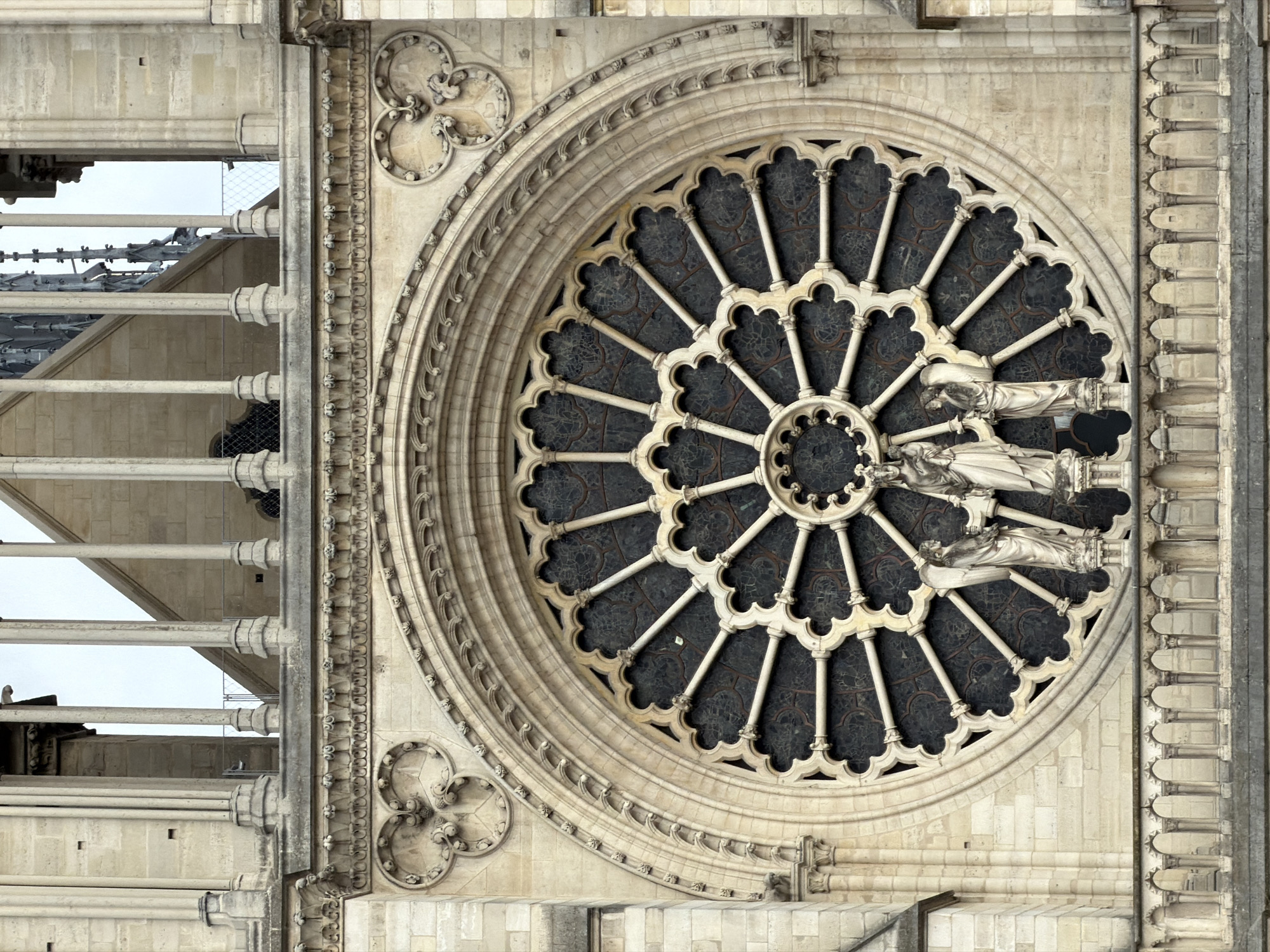 This media file is a close-up, high-angle shot of a segment of a Gothic cathedral's facade, likely Notre Dame Cathedral in Paris, France. The primary subject is a large, ornate rose window, characterized by intricate stone tracery radiating from a central point. The window is filled with dark, patterned glass. Small, white stone figures, appearing to be statues or carvings of people, are embedded within the stonework of the rose window, positioned along some of the radiating spokes.
To the left of the rose window, the vertical structure of the cathedral is visible, featuring a series of parallel, horizontal stone beams or buttresses and lighter-colored stone blocks. This section offers a glimpse of the building's architectural complexity and scale. The sky appears overcast, suggesting a daytime scene, possibly late afternoon or early morning given the diffuse light. The overall impression is one of grandeur, historical significance, and meticulous craftsmanship. There is no visible text in the image.