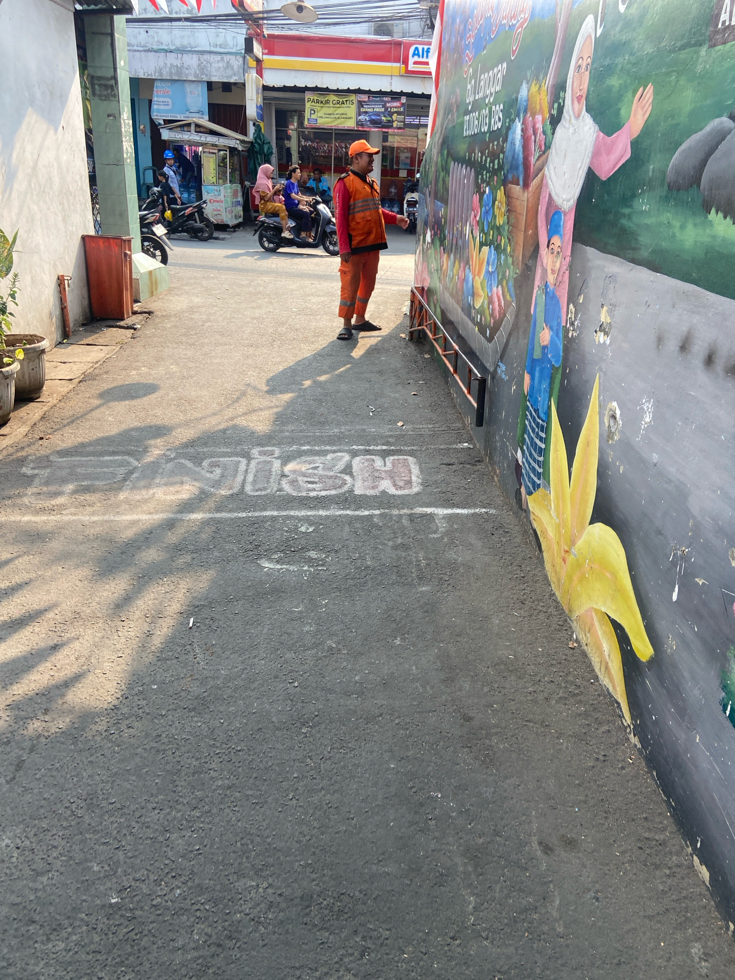 This is a medium shot, eye-level, outdoor, full-color photograph taken in an alleyway in North Jakarta, Indonesia. The photograph captures a street scene with a mural on the right side of the frame. A sanitation worker, dressed in an orange uniform and cap, stands in the middle of the alley, facing left. In the background, a few motorcycles and people are visible, along with some storefronts. The ground of the alley is paved with asphalt, and a white line is painted across it, marking a "Finish" line. The mural itself depicts various figures and colorful elements, including flowers. The lighting suggests it is daytime, with shadows cast on the ground from the left side of the frame.