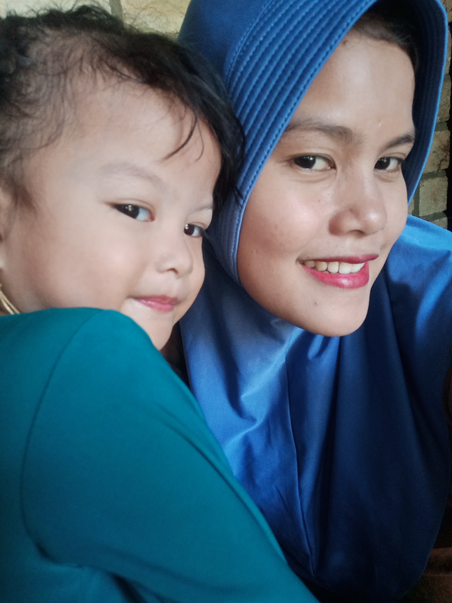 In a close-up, slightly upward-angled selfie, a woman wearing a bright blue hijab is seen with a child partially obscuring her left side. The woman's face is turned towards the camera, with her eyes looking directly at the viewer. She has a warm smile, revealing her teeth, and her lips are colored with bright pink lipstick. The child, on the left, is facing away from the camera, looking slightly to the left. The child has dark hair and is wearing a teal-colored garment. The background appears to be a brick wall, suggesting an indoor or possibly an outdoor setting with a brick structure. The lighting suggests a daytime photograph, possibly taken with natural light.