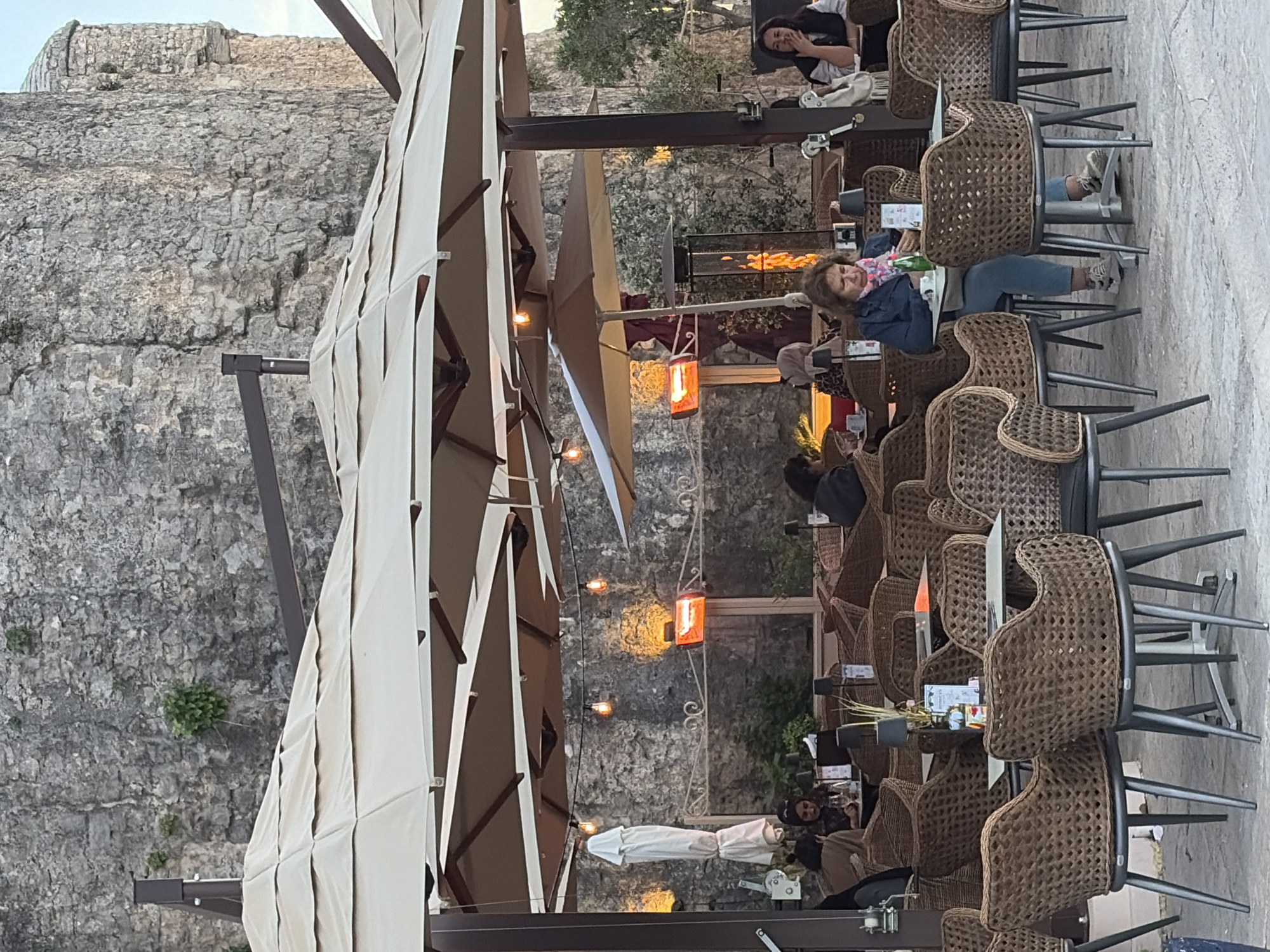 The image captures an outdoor cafe scene nestled against a towering, rugged stone wall, suggesting a historic or ancient setting like the Old Town of Budva, Montenegro. Large, cream-colored patio umbrellas extend over multiple tables, providing shade or cover.
Numerous wicker-style chairs with dark metal frames are arranged around square and rectangular tables, many of which are occupied by patrons. Several people are visible, engaged in conversation or simply relaxing. In the mid-ground, a woman with brown hair, wearing a denim jacket and a colorful scarf, is seated at a table with a green bottle, looking towards the viewer. Her feet are casually propped on an adjacent chair. Further back and to the right, another woman laughs, covering her mouth with her hand, suggesting a cheerful, social atmosphere. Other patrons are partially visible in the background and foreground, seated at various tables.
The scene is illuminated by warm ambient lighting from string lights strung between the umbrella structures and glowing orange patio heaters, indicating it is likely late afternoon or early evening. Menus are visible on some tables, though the specific text is illegible. The weathered stone wall, with its rough texture and occasional patches of greenery, forms a dramatic backdrop, contrasting with the modern cafe setup. The overall impression is one of a relaxed, inviting outdoor dining or lounge area in a unique historical environment.