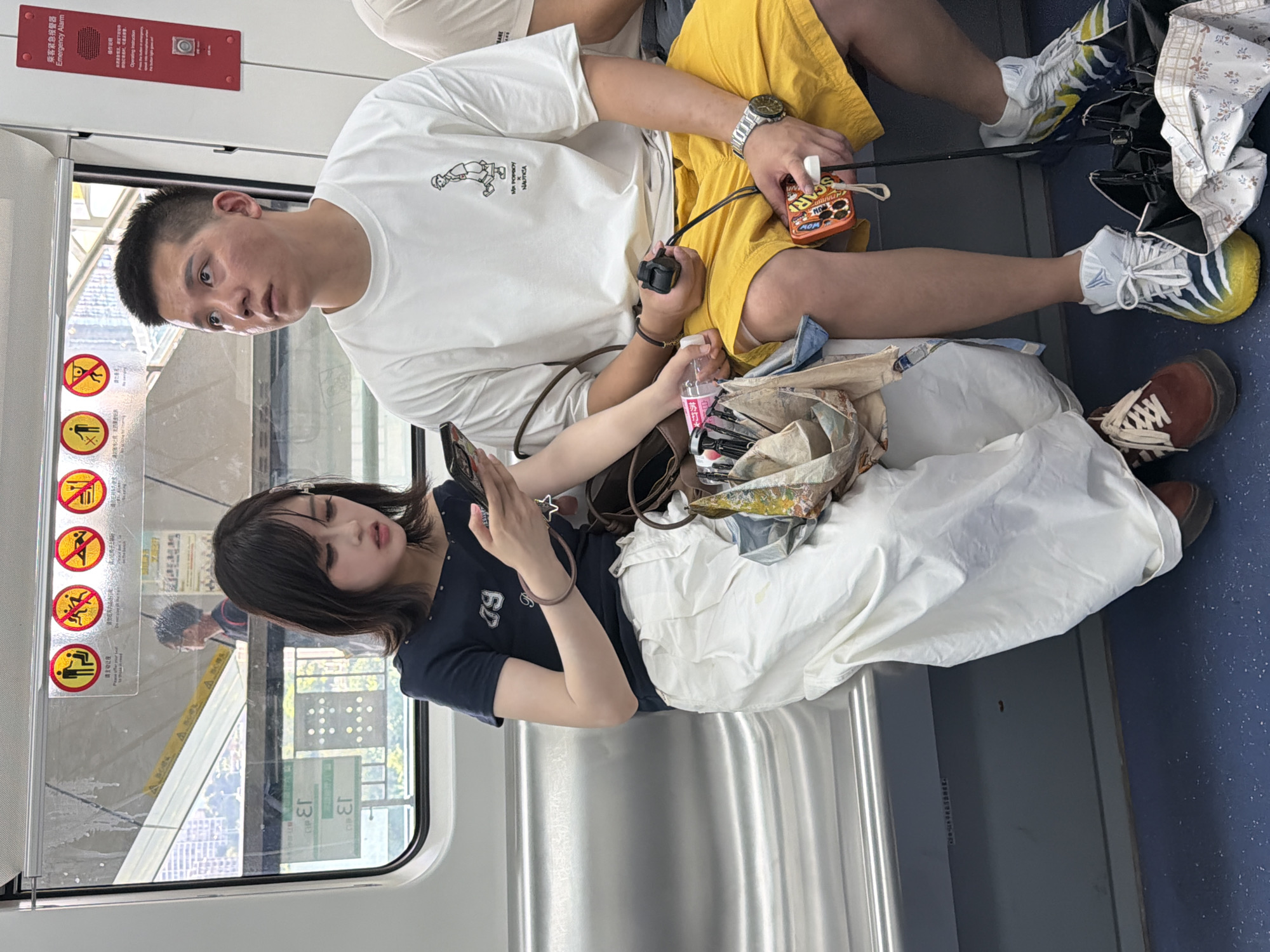 In the Yuzhong District of China, two individuals are seated on a metallic bench inside a public transportation vehicle. A young woman with dark hair, wearing a dark off-the-shoulder top and white wide-leg pants, is seated on the left. She is looking down at her smartphone, which she holds in her right hand. A brown star-shaped charm is attached to a strap near her hand. Next to her, on the right, is a man with short hair, wearing a white t-shirt with a graphic print and bright yellow shorts. He is looking off to his right. He holds a black remote-like object in his left hand and a small orange rectangular item, possibly a portable charger or case, in his right hand. Both individuals are wearing sneakers. The environment appears to be a train or metro car, with windows showing a blurry cityscape and posters displayed above the windows. The floor is a dark blue color.