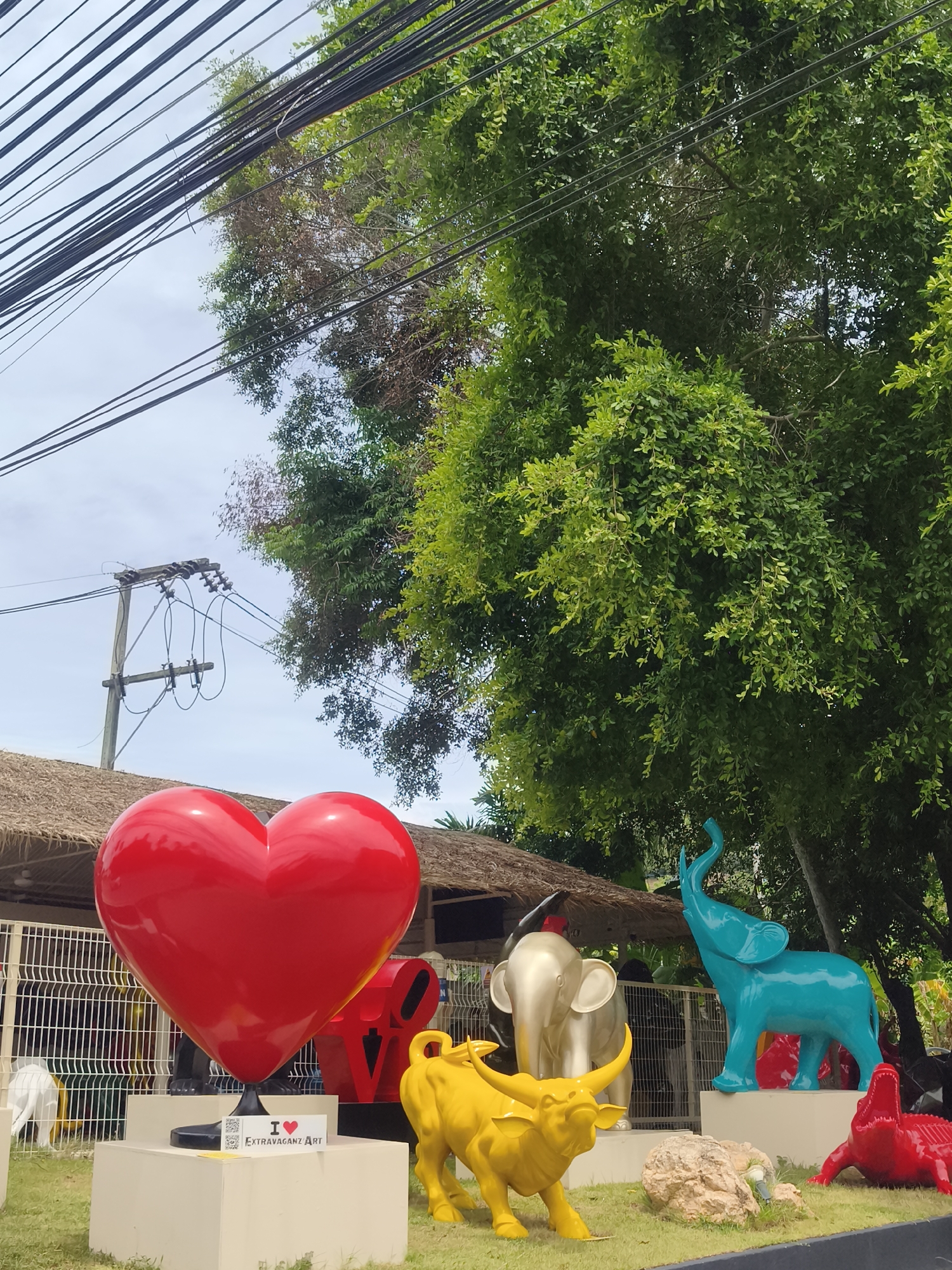 This is an outdoor scene in Ko Samui District, Thailand, with a focus on a collection of colorful animal sculptures and a large red heart. In the foreground, there is a bright yellow bull sculpture and a red crocodile sculpture. Behind them, a large teal elephant sculpture is positioned on a pedestal. To the left, a large, glossy red heart sculpture stands on a white pedestal, with a QR code and "I❤️ ExtraVaganz Art" sign at its base. In the middle ground, a thatched-roof structure is visible, behind a white fence. Overhead, a dense canopy of green trees dominates the upper portion of the image, with a network of black electrical wires crisscrossing in front of a pale sky. The overall environment appears to be a public space, possibly an art installation or an outdoor exhibition area.