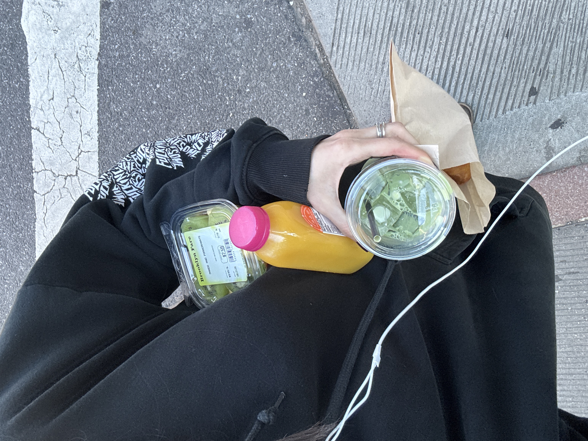 The image shows a person sitting on a city sidewalk, likely in New York City, as indicated by the "Brooklyn Fare" label on a food container. The person is wearing a black hoodie with a graphic print on the sleeve and black pants. They are holding several items: a clear plastic container filled with sliced kiwi, a yellow bottle with a pink cap filled with juice, and a clear plastic cup containing a green beverage with ice. A brown paper bag is also visible, partially concealing a pastry. White earbuds are plugged into the person's ear and draped across their lap. The background consists of a gray asphalt road with a white crosswalk stripe, suggesting an urban street environment. The lighting indicates it is daytime.