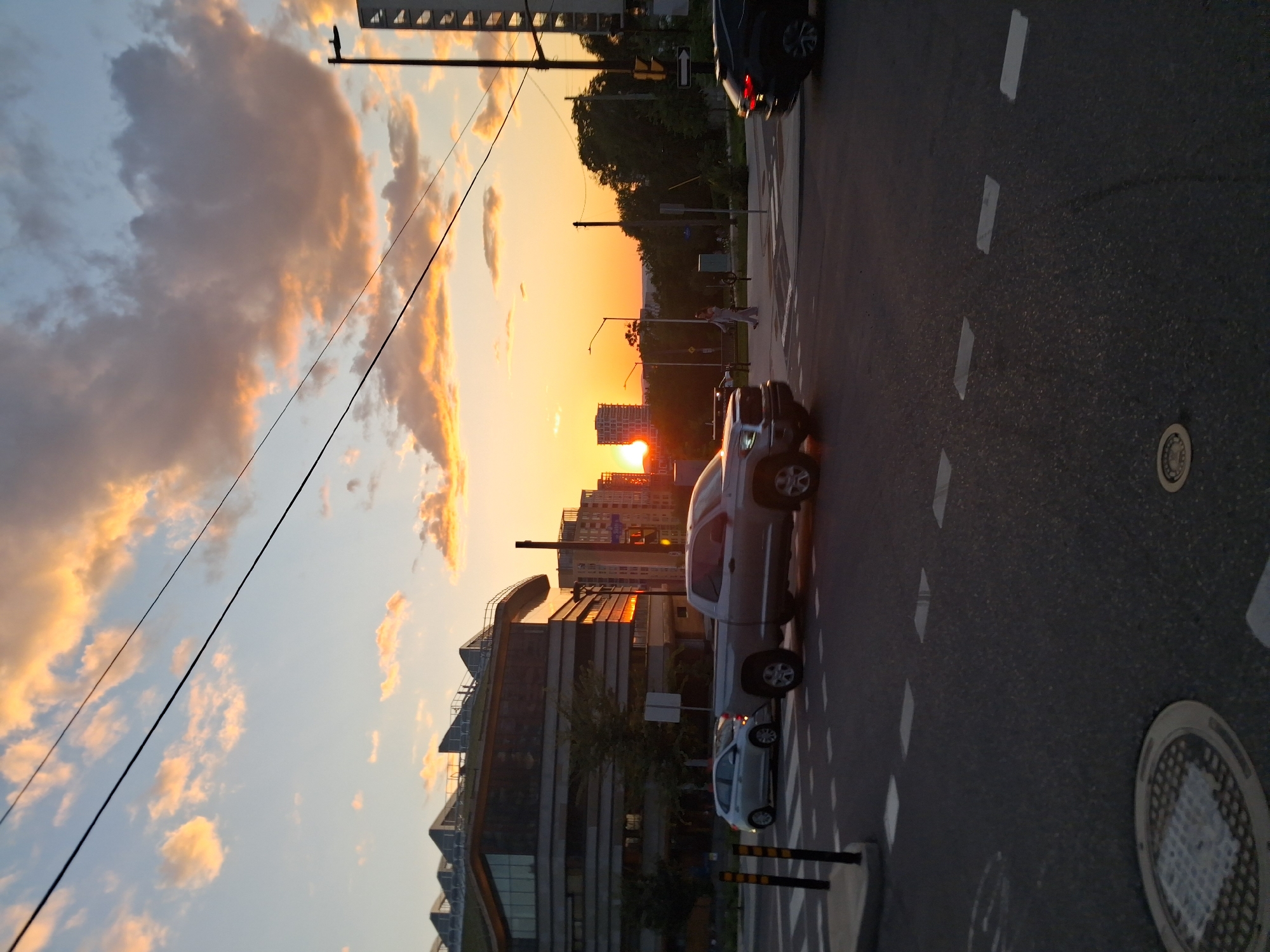 The image captures a street scene at sunset in Ottawa, Canada. A silver pickup truck and a silver sedan are at an intersection with crosswalk markings. In the background, modern buildings with distinctive angled roofs and glass facades are visible. The sky is filled with clouds tinged orange and yellow by the setting sun. A street light stands on the right side of the image. Further in the distance, more buildings and trees are silhouetted against the vibrant sky. Two pedestrians are walking on the sidewalk. The foreground shows the asphalt road with visible manhole covers. The image appears to be taken from a street-level perspective.