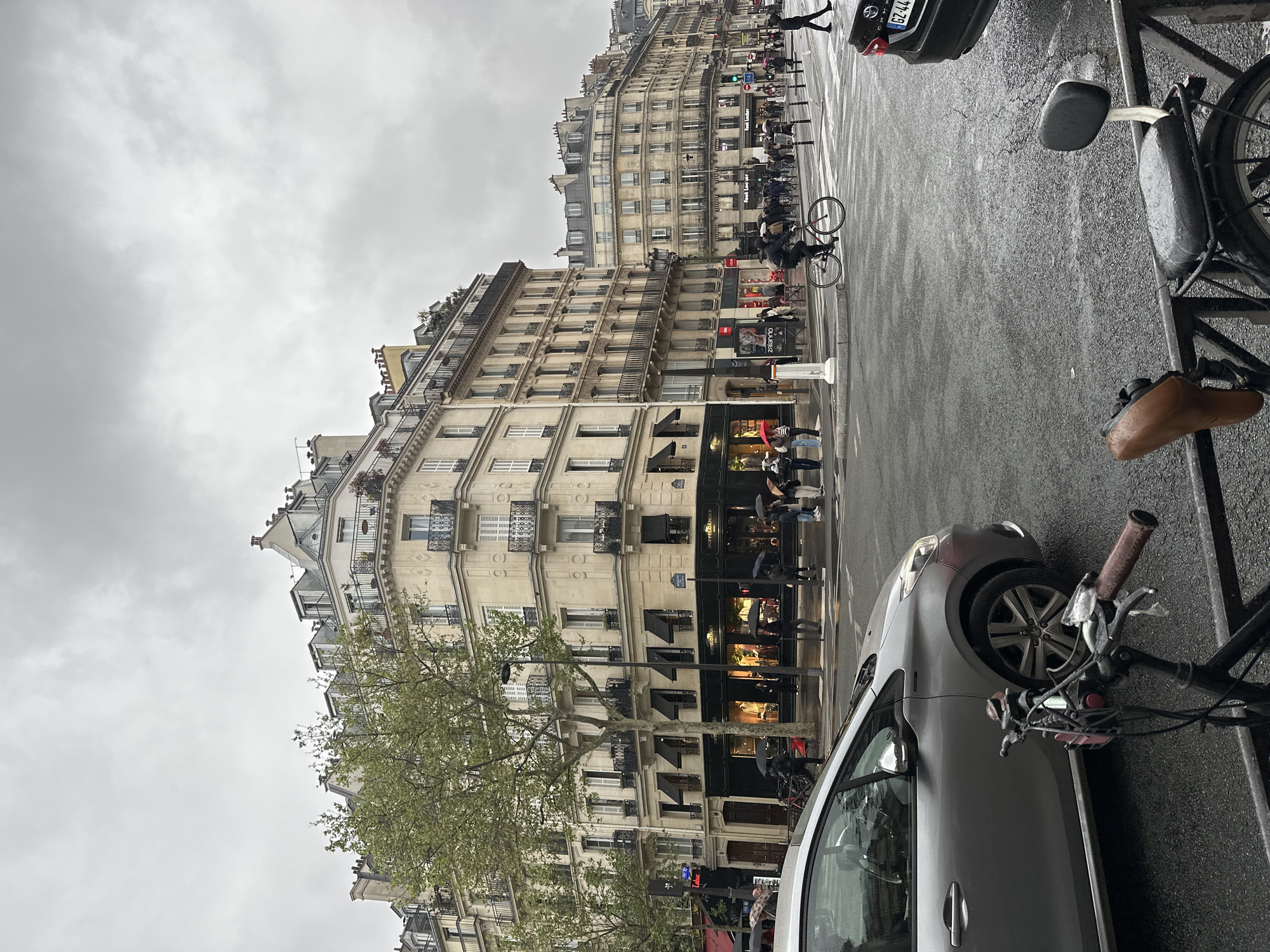 The image shows a street scene in Paris, France, on a rainy day. Pedestrians with umbrellas are crossing the street. Buildings with multiple floors and classic Parisian architecture line the street. A bicycle is parked in the foreground and a car is visible. The sky is overcast.