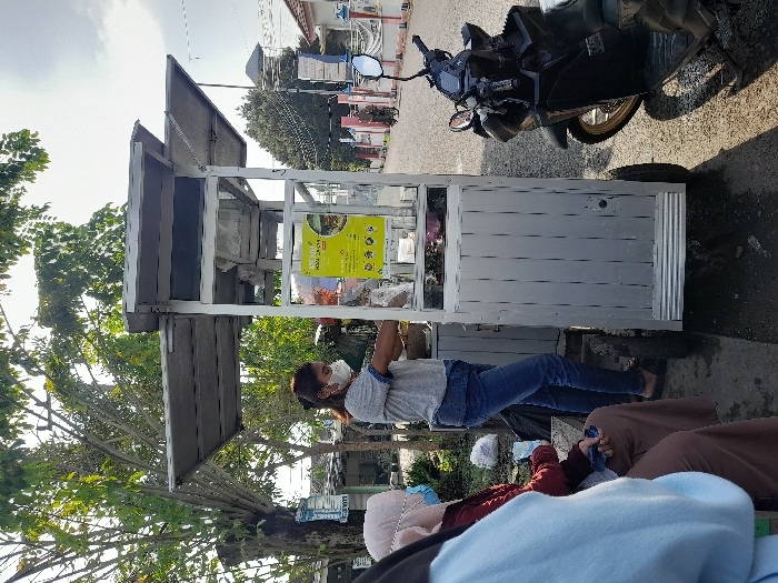 A sideways view shows a woman in a mask standing beside a food cart. She wears a grey shirt and jeans. The cart is light grey with a silver frame, displaying a yellow poster. A black scooter is parked nearby on the paved street. Several people are seated near the bottom of the frame, partially obscured, wearing various colors. Buildings and trees are visible in the background under a bright sky.
