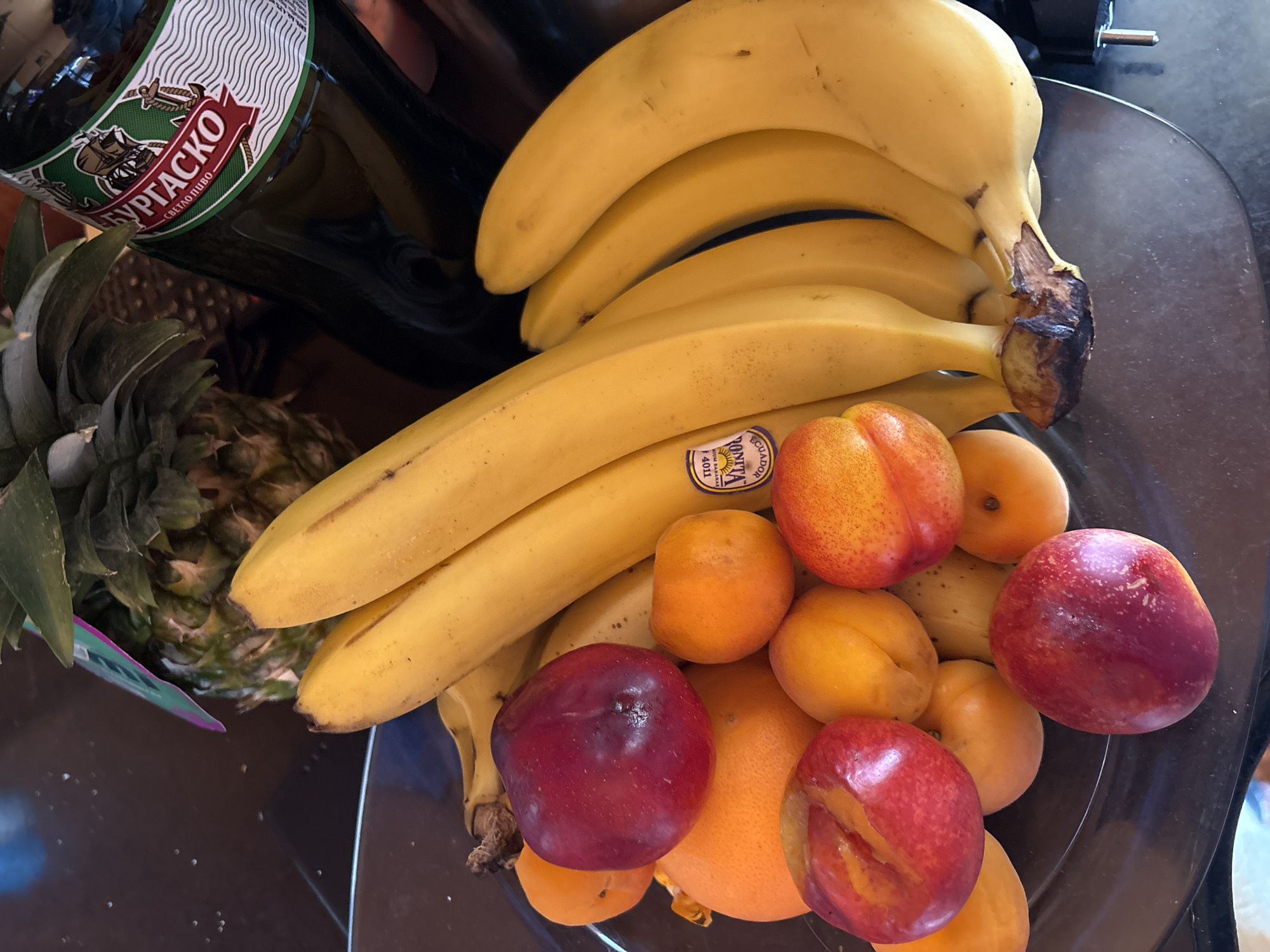 A close-up shot showcases a bowl of fruit in Unknown, Unknown. The composition includes a bunch of ripe bananas, several apricots, and three plums. A pineapple and a beer bottle, labeled "Бургаско" (Burgasko), are partially visible on the side. The arrangement sits on a dark surface.